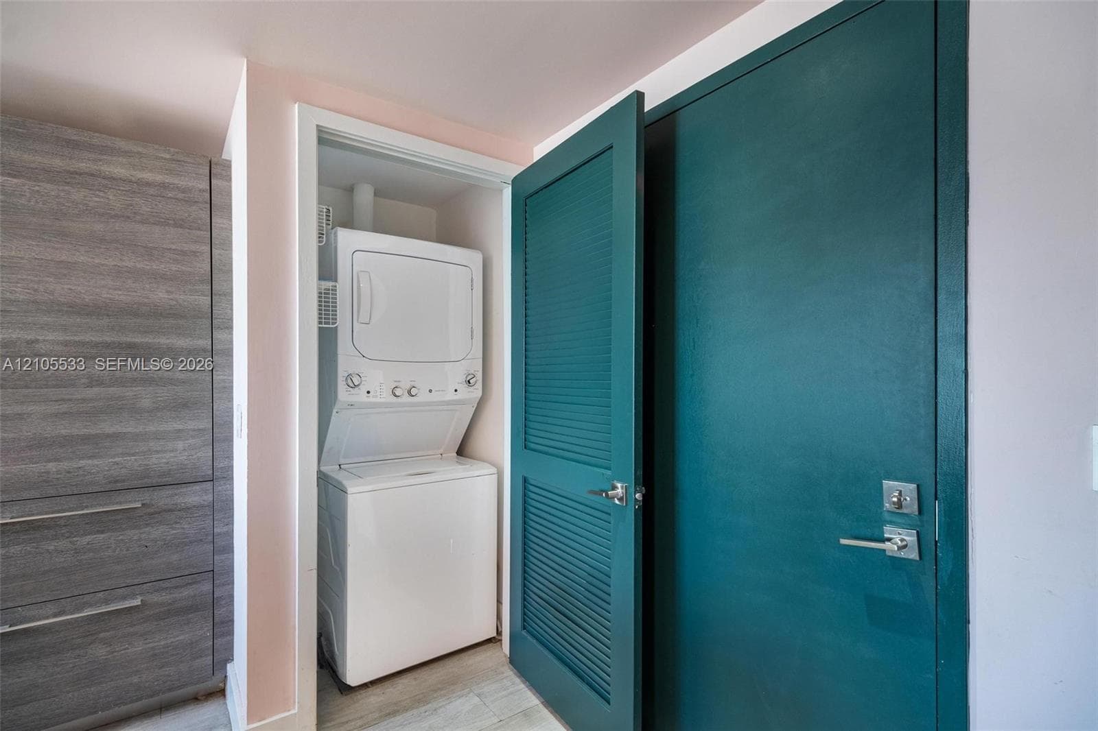 Stacked laundry unit housed in a dedicated closet, featuring a teal louvered door, wood-finish built-in cabinetry, and wood-finish flooring