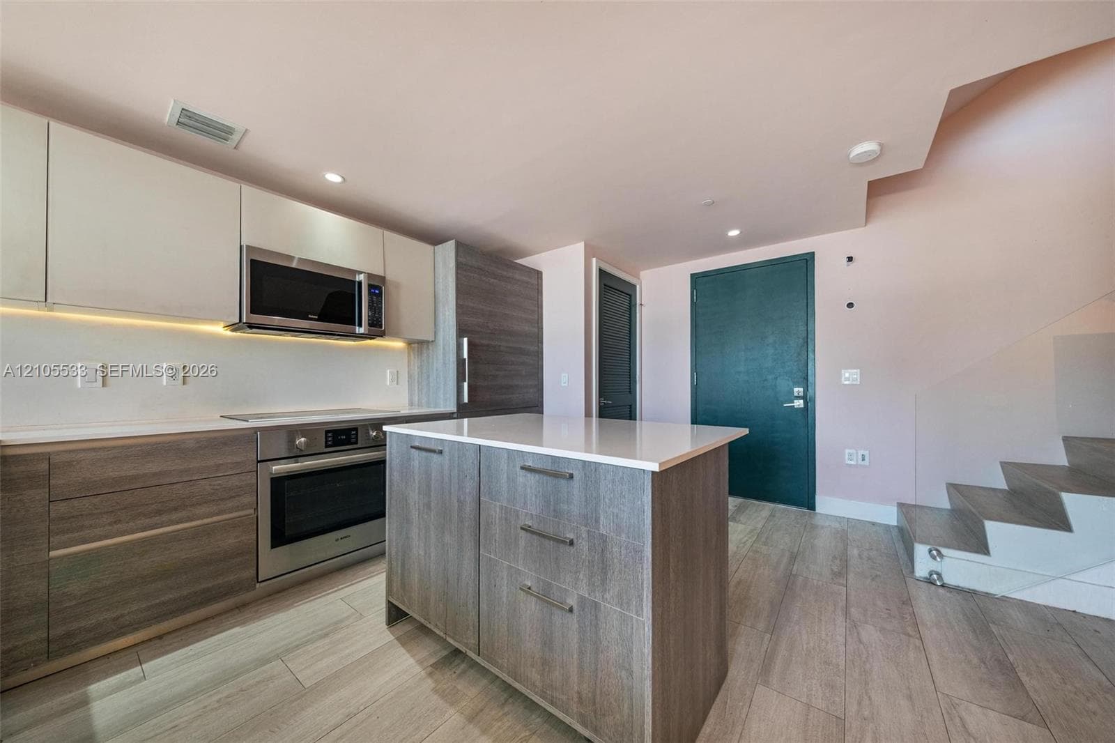 Kitchen featuring flat-panel cabinetry, a central island with a solid-surface countertop, stainless steel appliances, wood-finish flooring, and a glass-paneled staircase