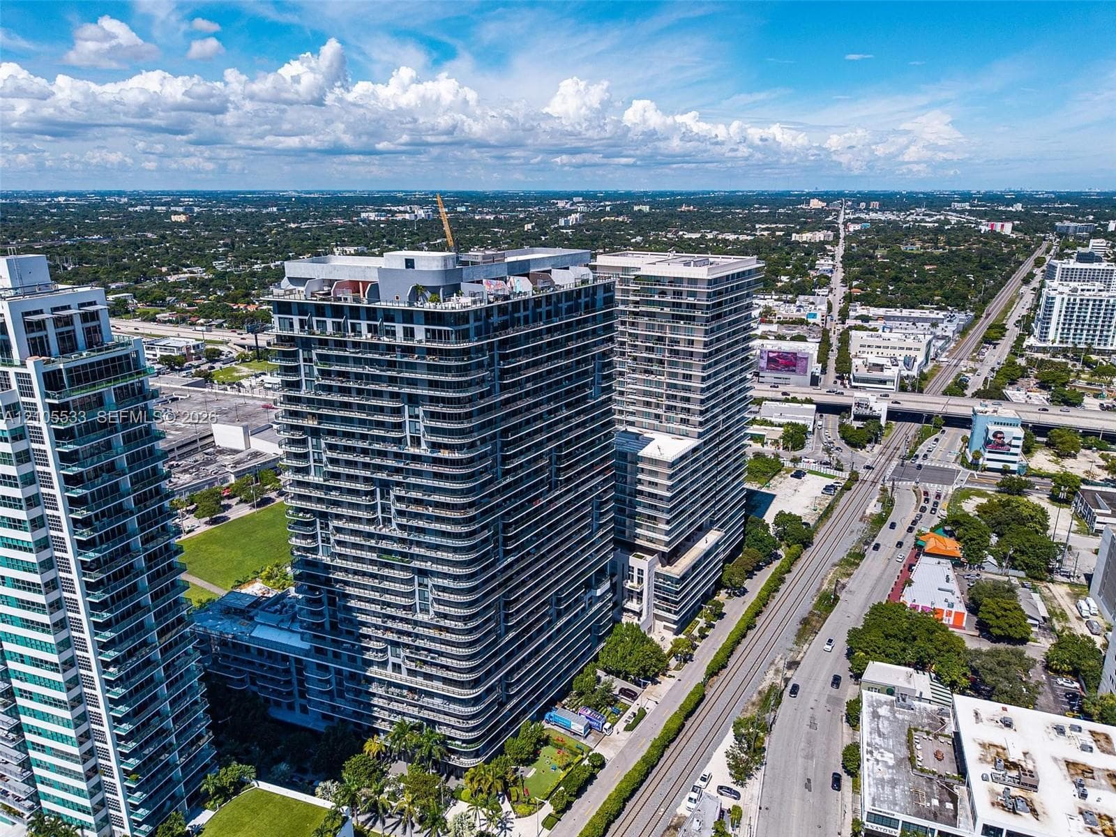 High-rise residential towers featuring wrap-around balconies, glass-paneled railings, exterior concrete structural elements, rooftop recreational space, and surrounding landscaped grounds