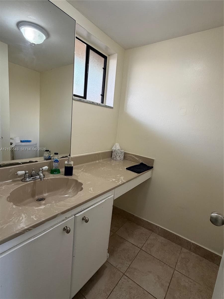 Bathroom featuring a vanity with a solid-surface countertop, integrated basin, large wall-mounted mirror, ceramic tile flooring, and a window with privacy glass