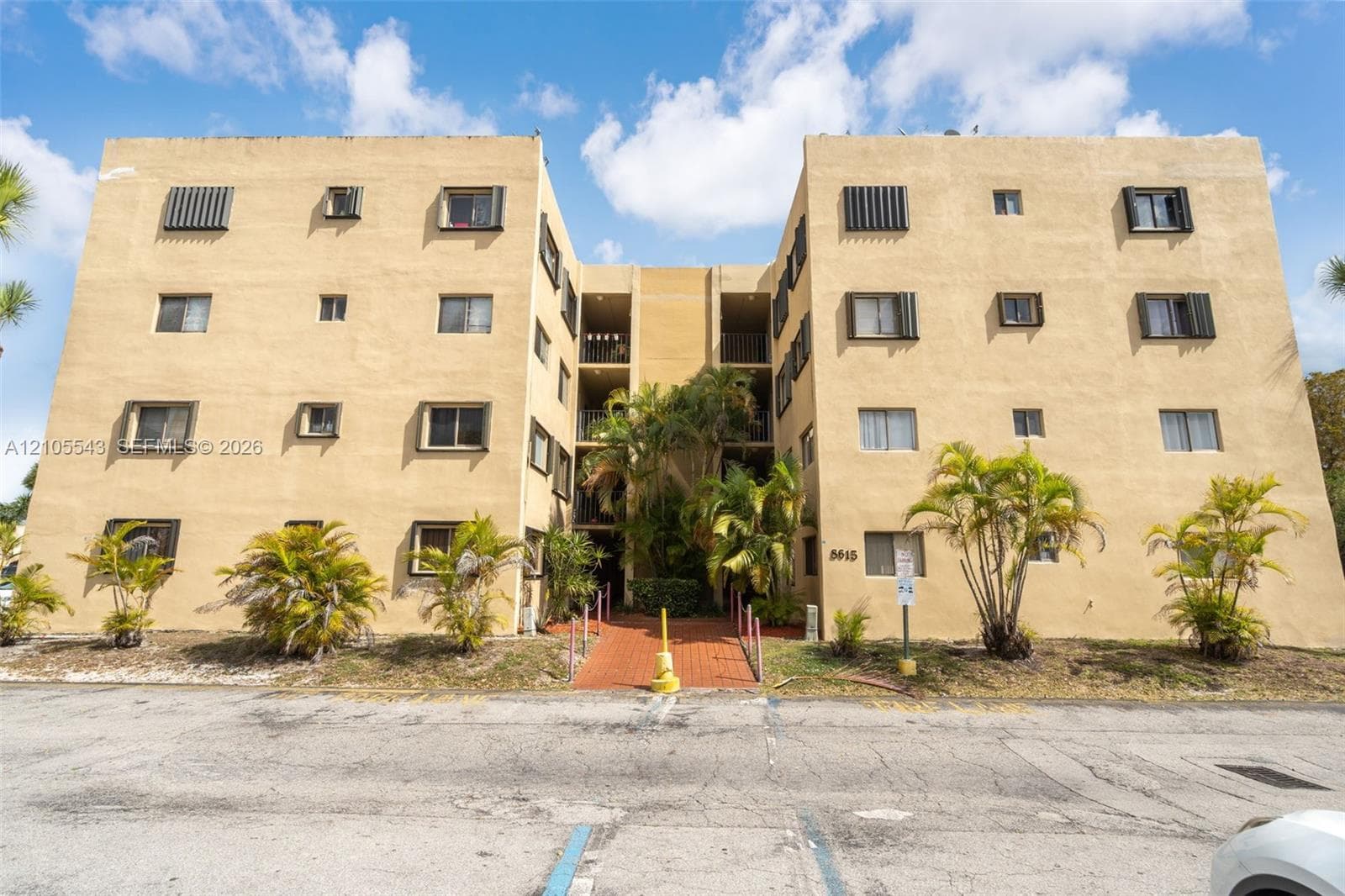 Multi-story exterior featuring a stucco facade, integrated balcony access, red brick paver walkway, and surrounding palm trees