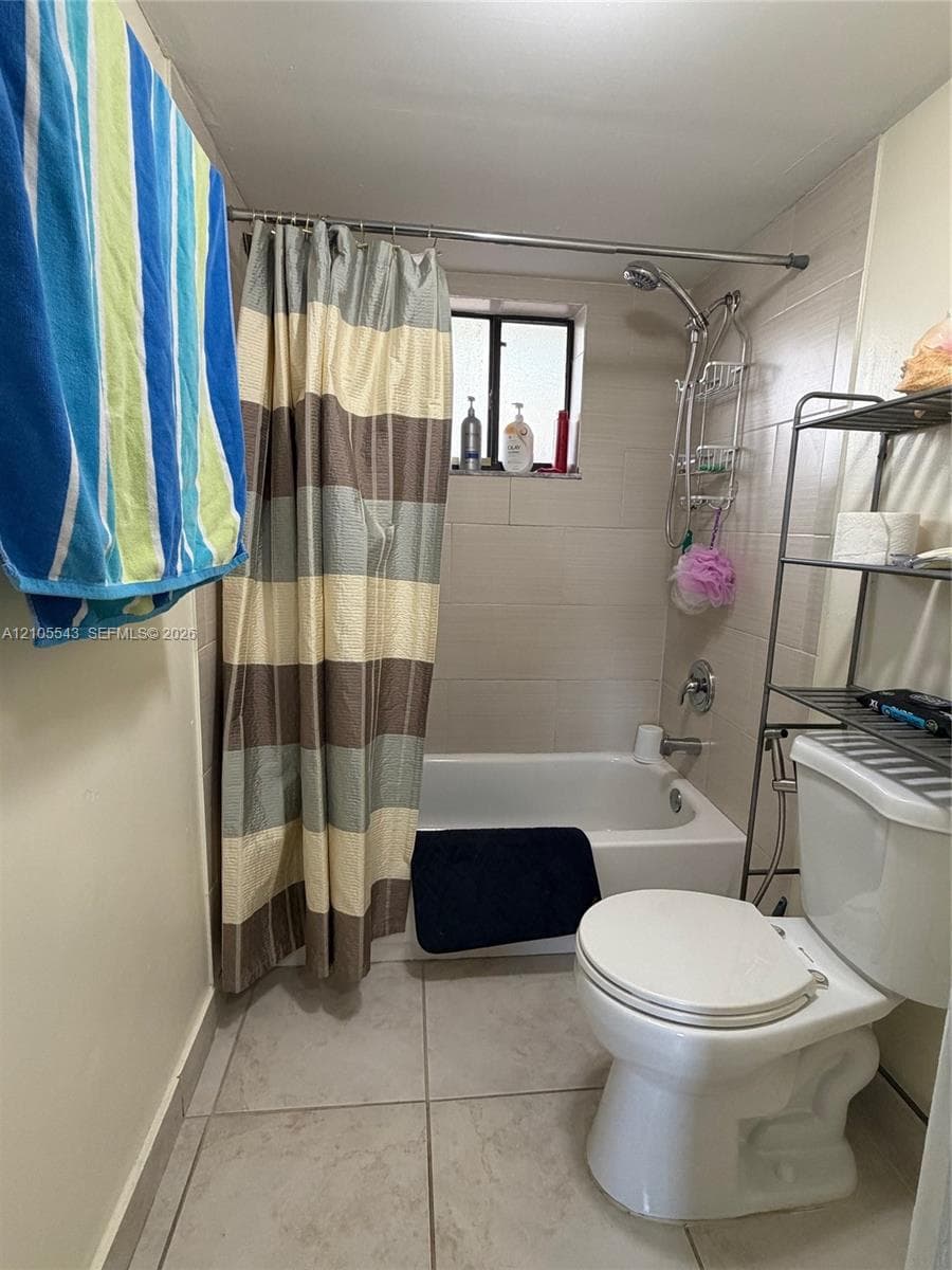 Bathroom featuring a white porcelain tub-shower combination, wall-mounted shower head, neutral-toned ceramic wall tile, and light-colored floor tiling