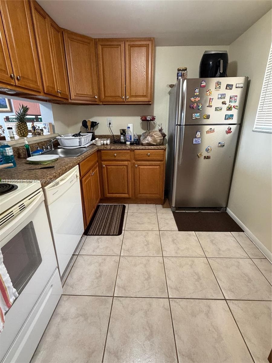Kitchen featuring wood cabinetry, solid surface countertops, stainless steel refrigerator, white dishwasher, and neutral ceramic tile flooring