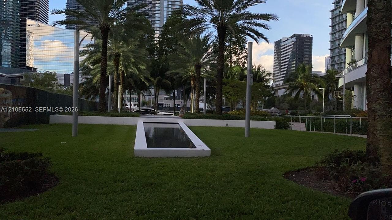 Expansive green space featuring a decorative water element, mature palm trees, contemporary lighting fixtures, and high-rise structures in the background