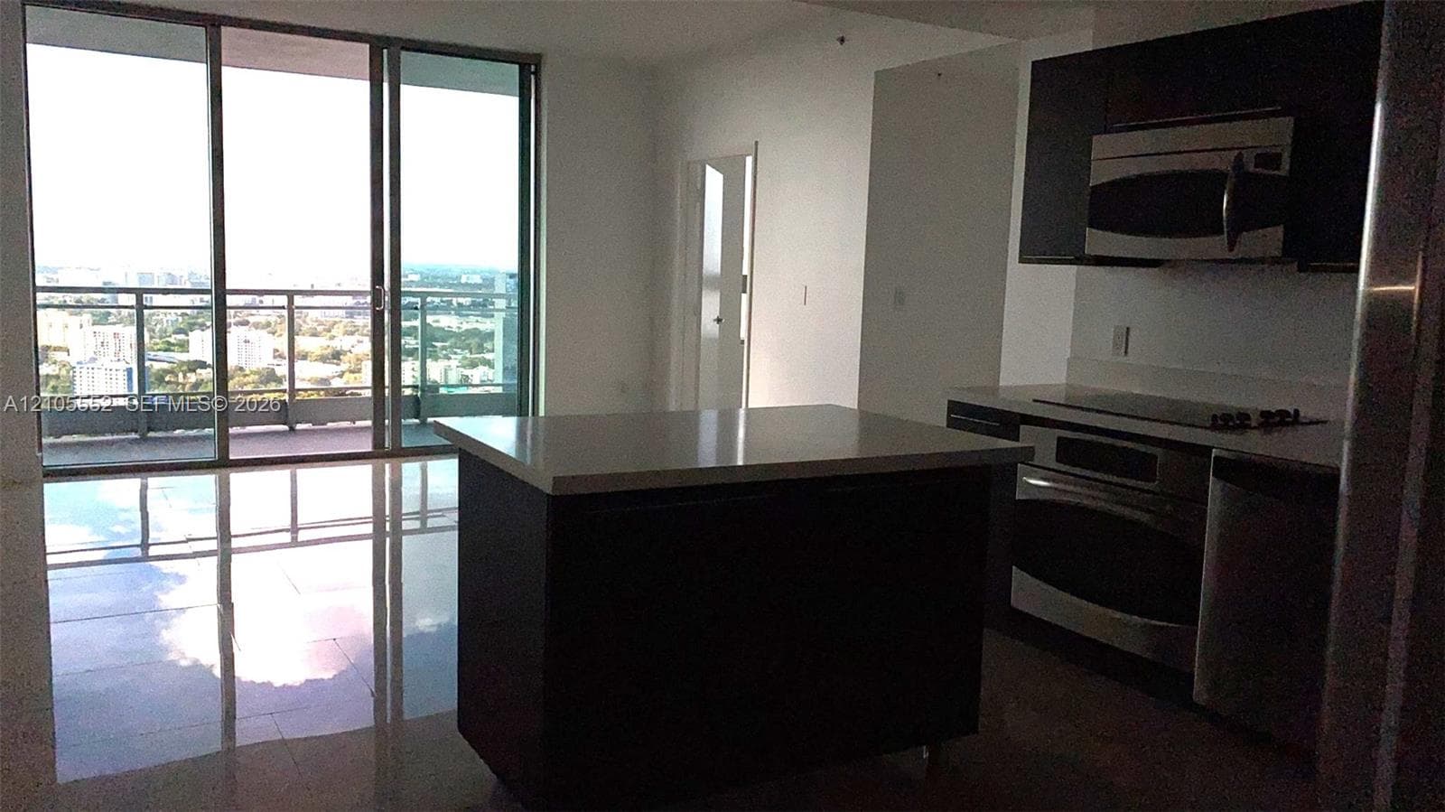 Open-concept living space with large sliding glass doors, a kitchen island with a light-toned countertop, dark wood-finish cabinetry, and stainless steel appliances