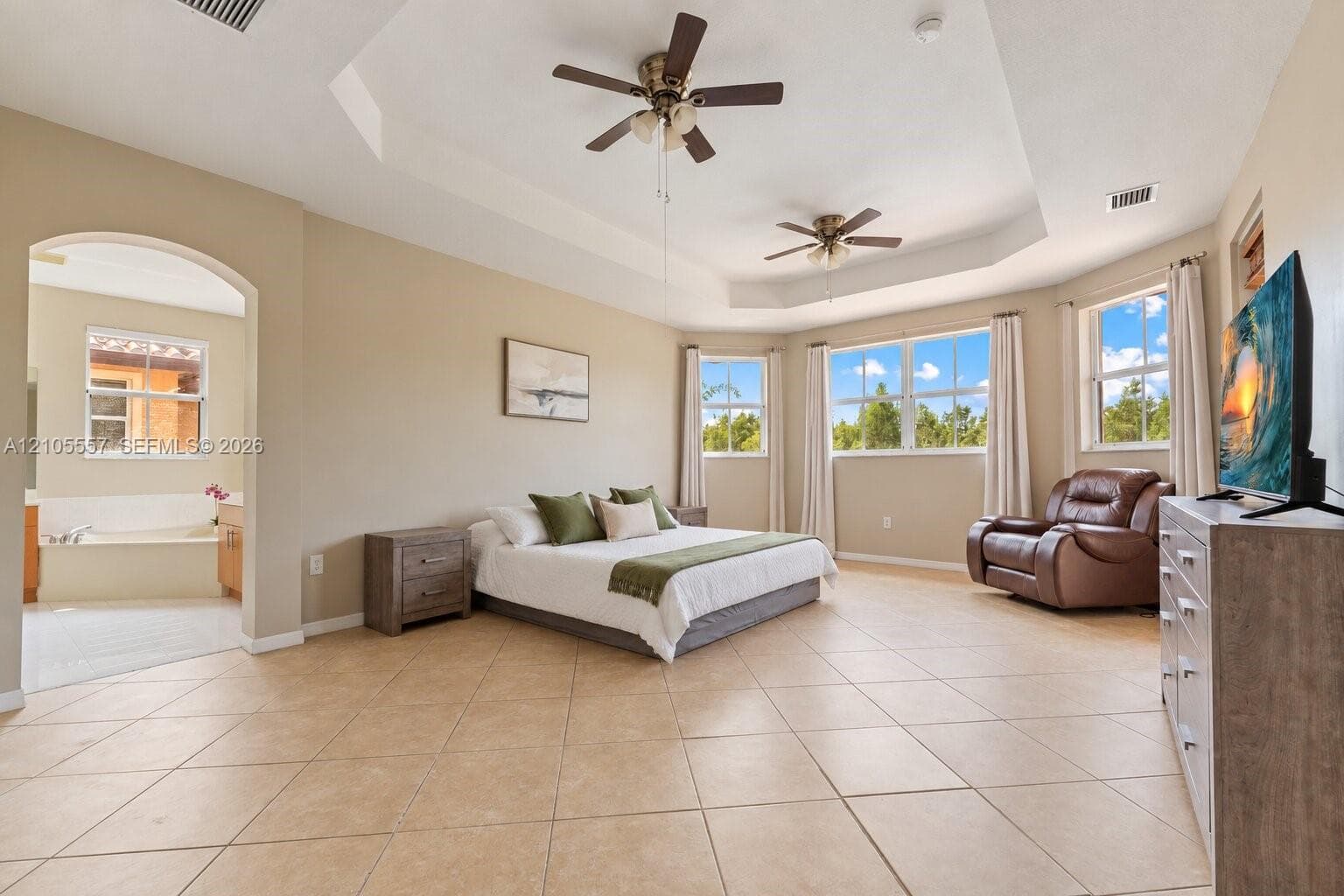 Tray ceiling with two ceiling fans, tile flooring, multiple windows, and an arched entry to an additional space with a built-in tub