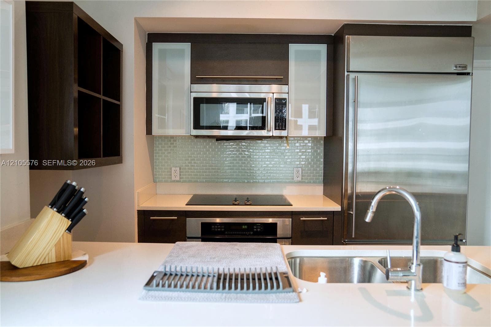 Kitchen featuring dark wood cabinetry, glass-front upper cabinets, stainless steel appliances, light-toned solid surface countertops, and a glass tile backsplash