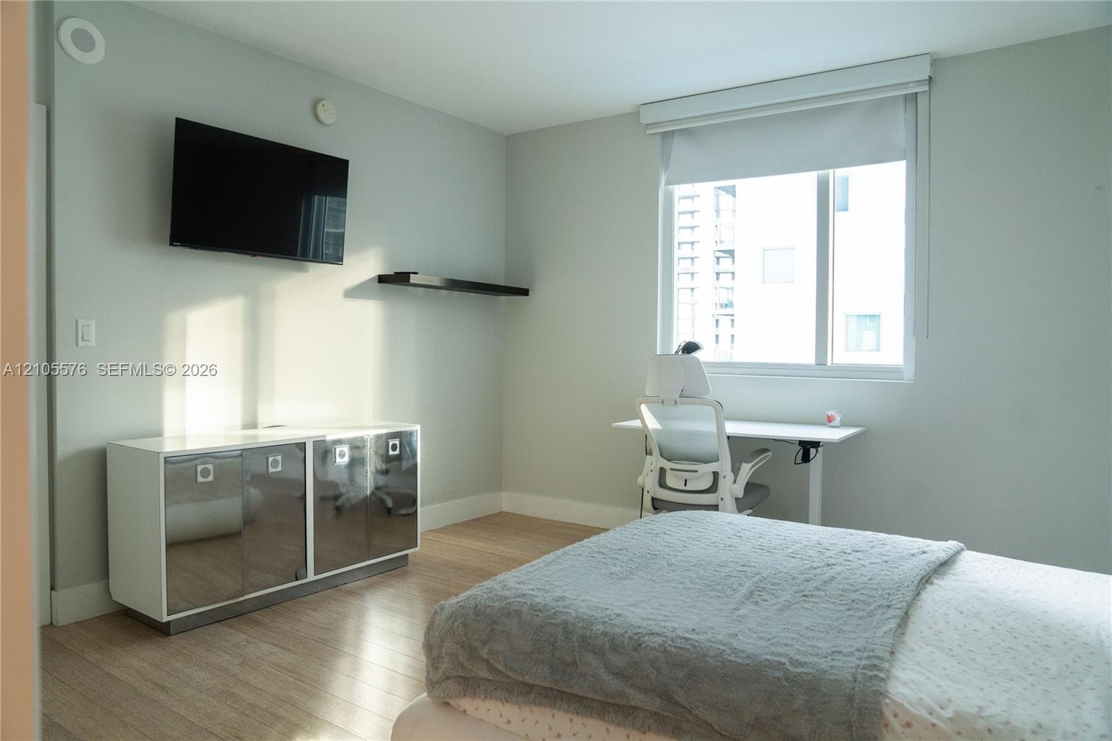 Room featuring wood-finish flooring, a wall-mounted floating storage unit with reflective finish, a fixed wall-mounted shelf, and a large window with roller shade
