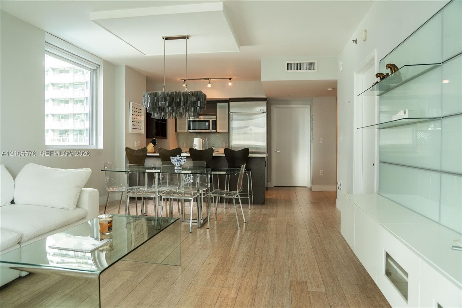 Open-concept interior featuring light wood-finish flooring, a contemporary island kitchen, built-in frosted glass shelving, and a decorative drop-pendant light fixture