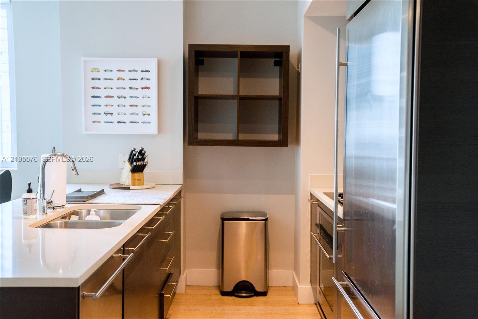 Kitchen featuring flat-panel dark cabinetry, white countertops, integrated stainless steel refrigerator, undermount double-basin sink with a gooseneck faucet, and wall-mounted grid shelving