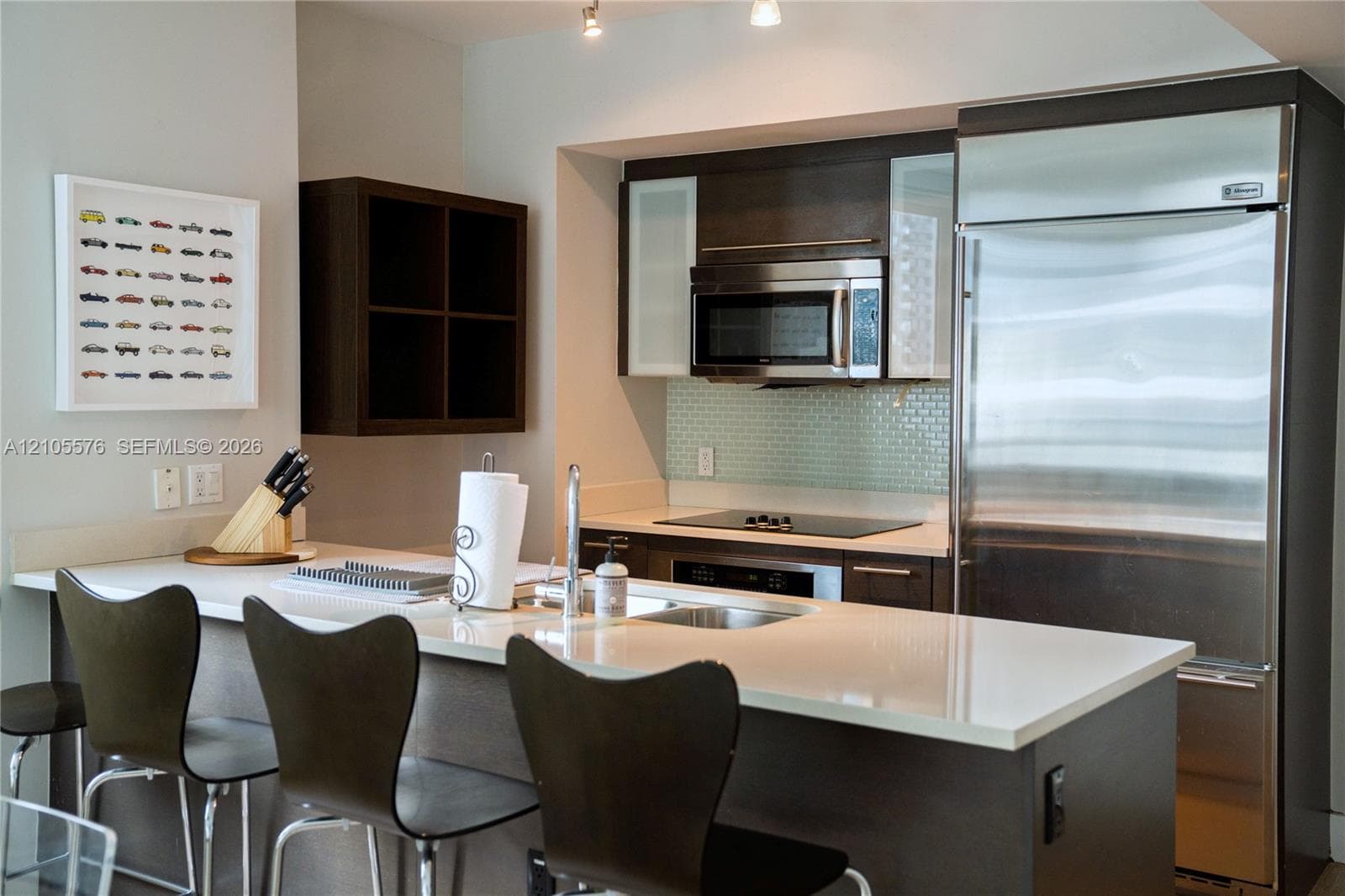 Kitchen featuring dark wood-finish cabinetry, light-toned stone countertops, a glass tile backsplash, stainless steel appliances, and an integrated breakfast bar