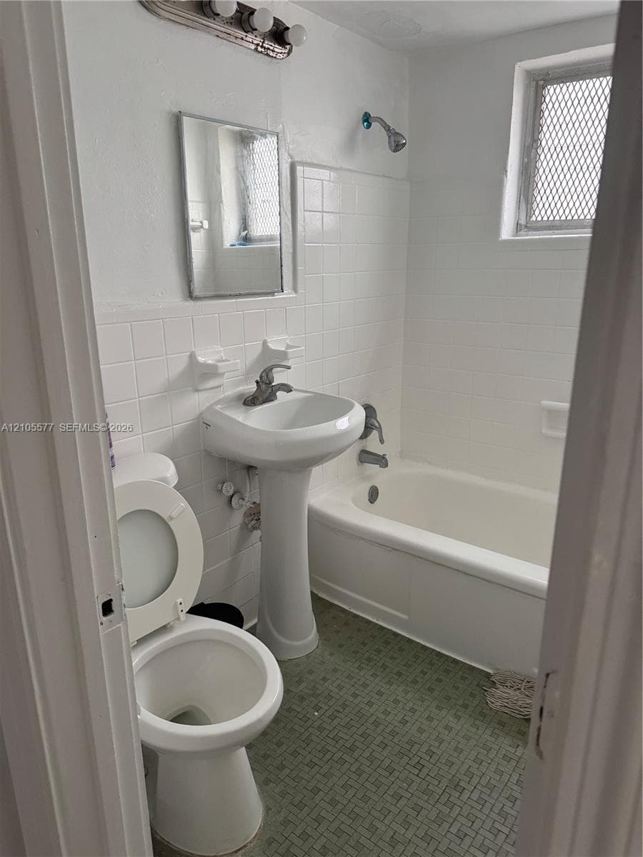 Bathroom featuring a white porcelain pedestal sink, white tiled tub surround, standard bathtub, integrated wall-mounted lighting, and textured flooring