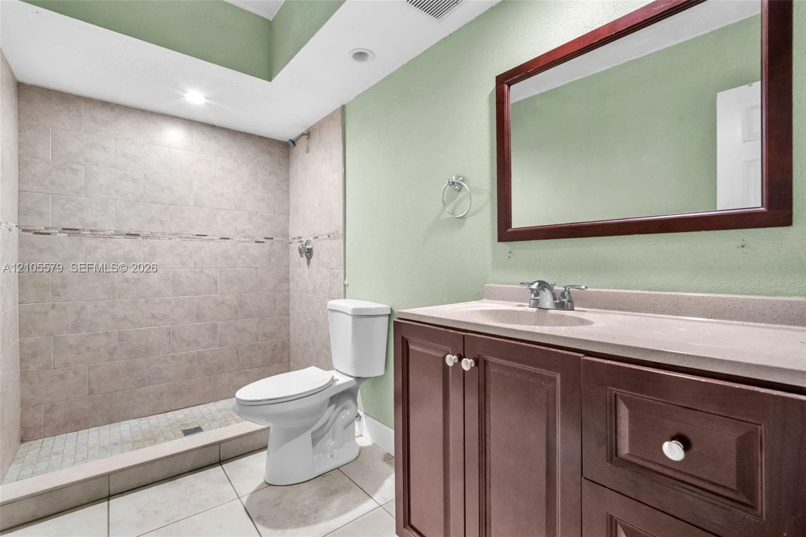 Bathroom featuring a walk-in tiled shower with an accent border, vanity with integrated stone-finish countertop, dark wood-finish cabinetry, and a framed wall-mounted mirror