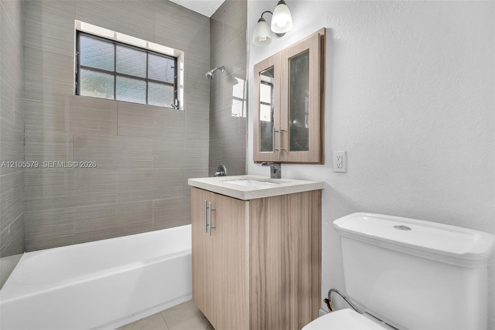 Bathroom featuring a tile-surround bathtub with a rectangular window, light wood-finish vanity cabinet, stone-composite countertop, and a double-door medicine cabinet