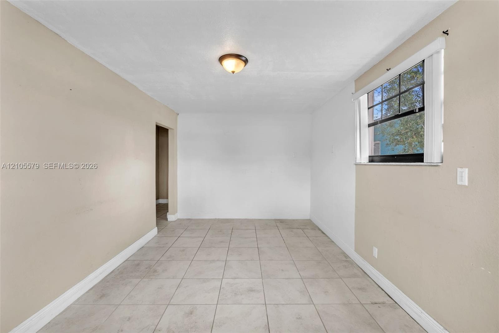 Neutral-toned interior space featuring light-colored tile flooring, smooth walls, a single-pane window with dark muntins, and a flush-mount ceiling light fixture