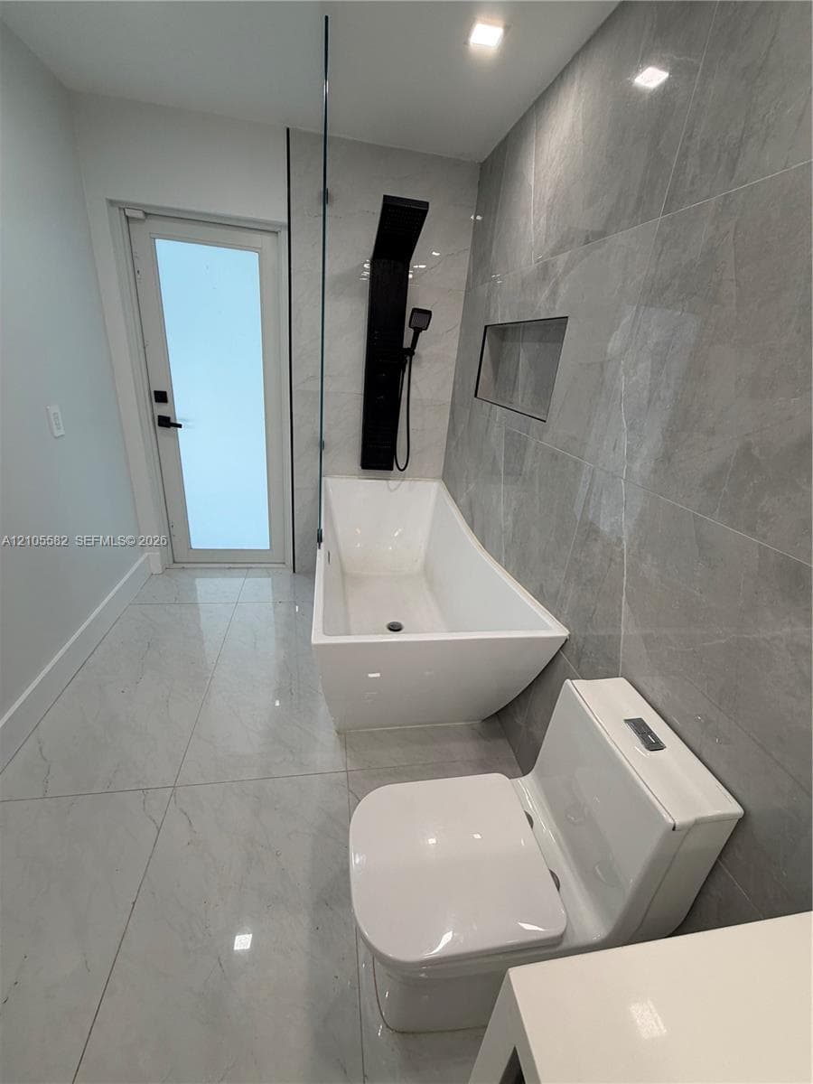 Floor-to-ceiling gray-veined tile, freestanding soaking tub with black shower panel, integrated wall niche, white toilet, and recessed lighting