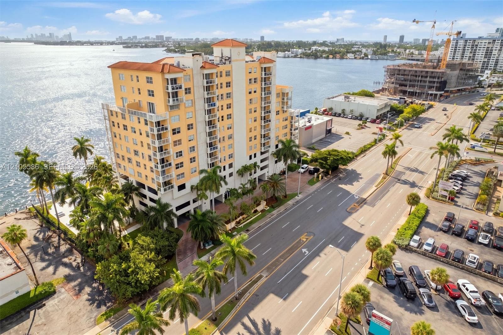Multi-story residential building with private balconies overlooking a body of water, tropical landscaping, and an adjacent multi-lane road