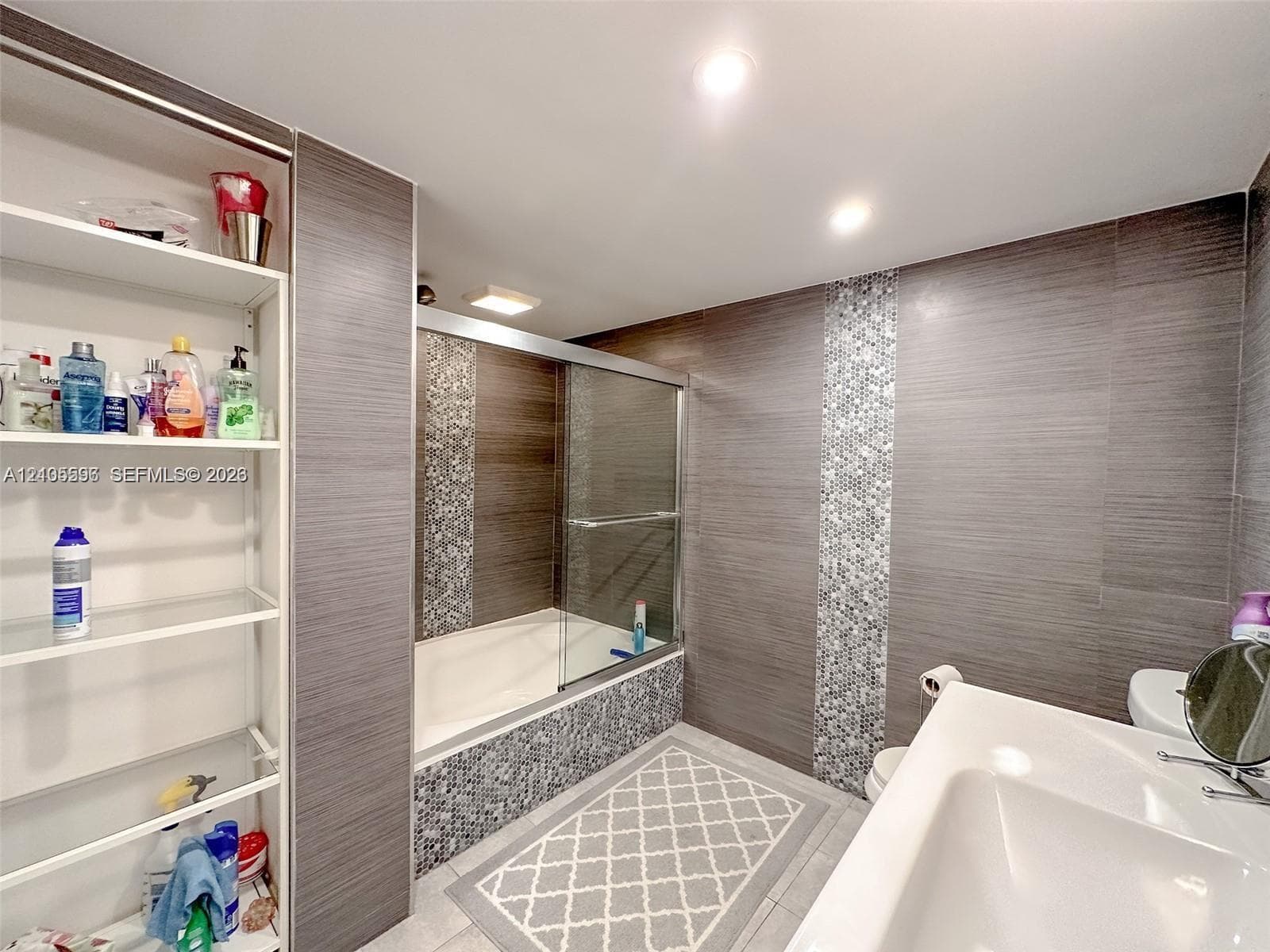 Bathroom featuring large-format horizontal gray wall tiling, vertical glass mosaic tile accents, a built-in recessed shelving unit, a glass-enclosed bathtub, and recessed ceiling lighting