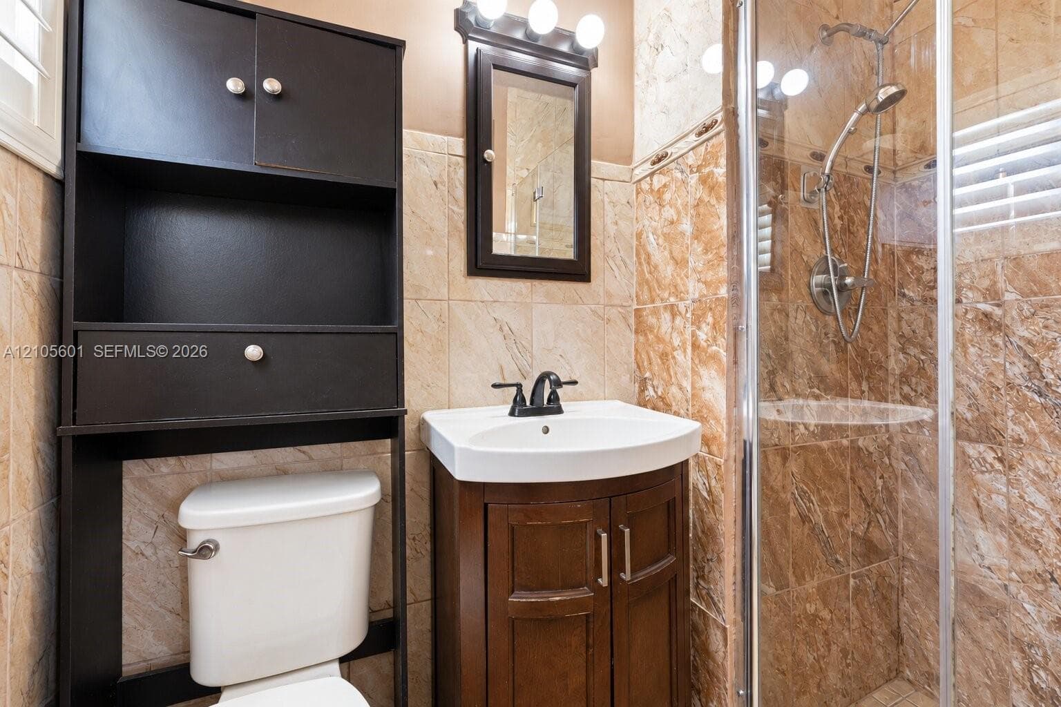 Glass-enclosed shower with a handheld shower fixture, wood-finish vanity with a white basin, dark wood-finish medicine cabinet, and tiled walls
