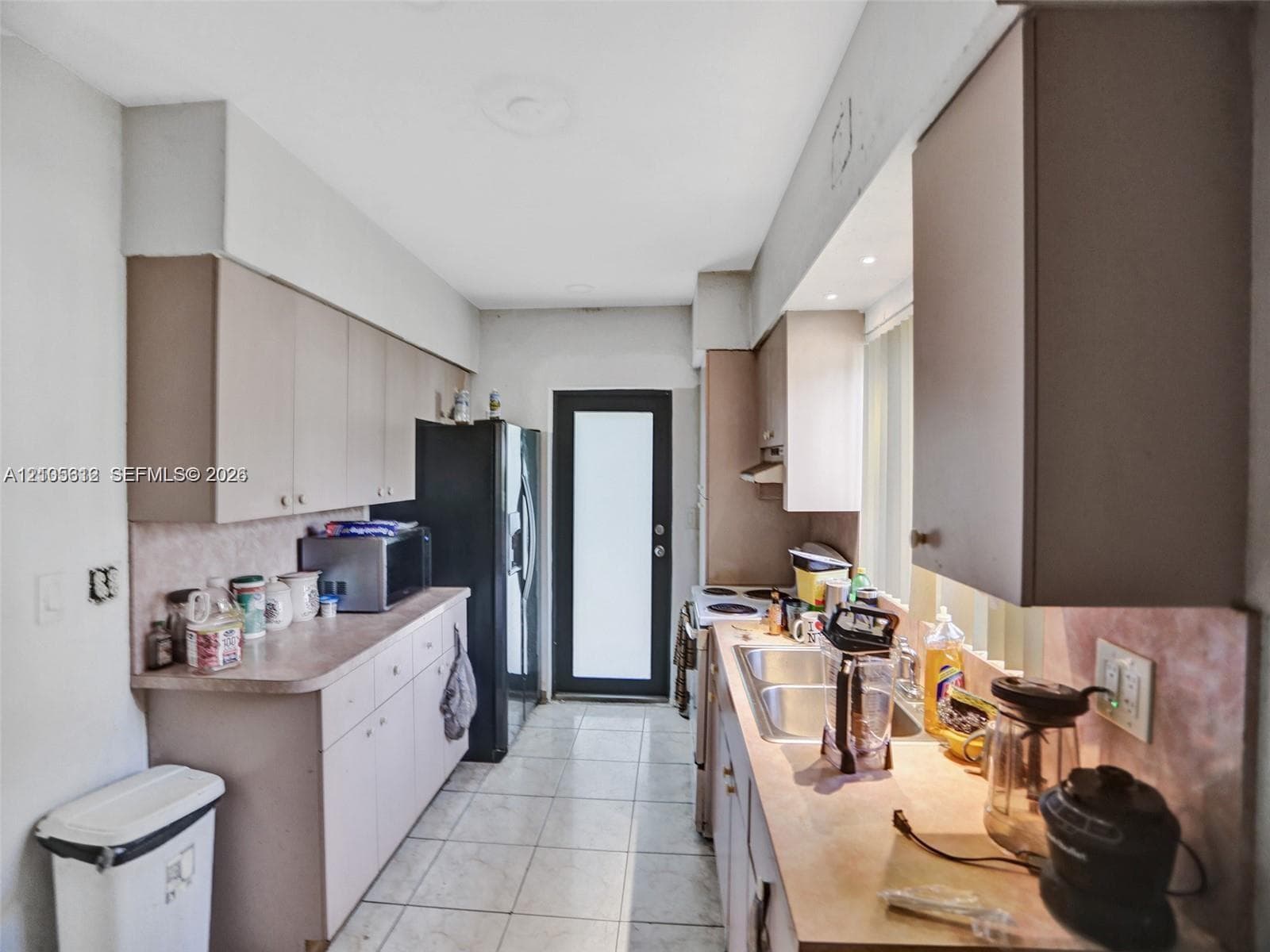 Galley-style kitchen featuring neutral-tone cabinetry, tiled flooring, solid-surface countertops, dual-basin stainless steel sink, and a glass-panel exterior door