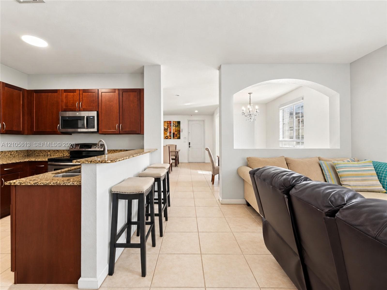 Kitchen featuring wood cabinetry, stone countertops, and an integrated breakfast bar, characterized by tile flooring and an arched pass-through window