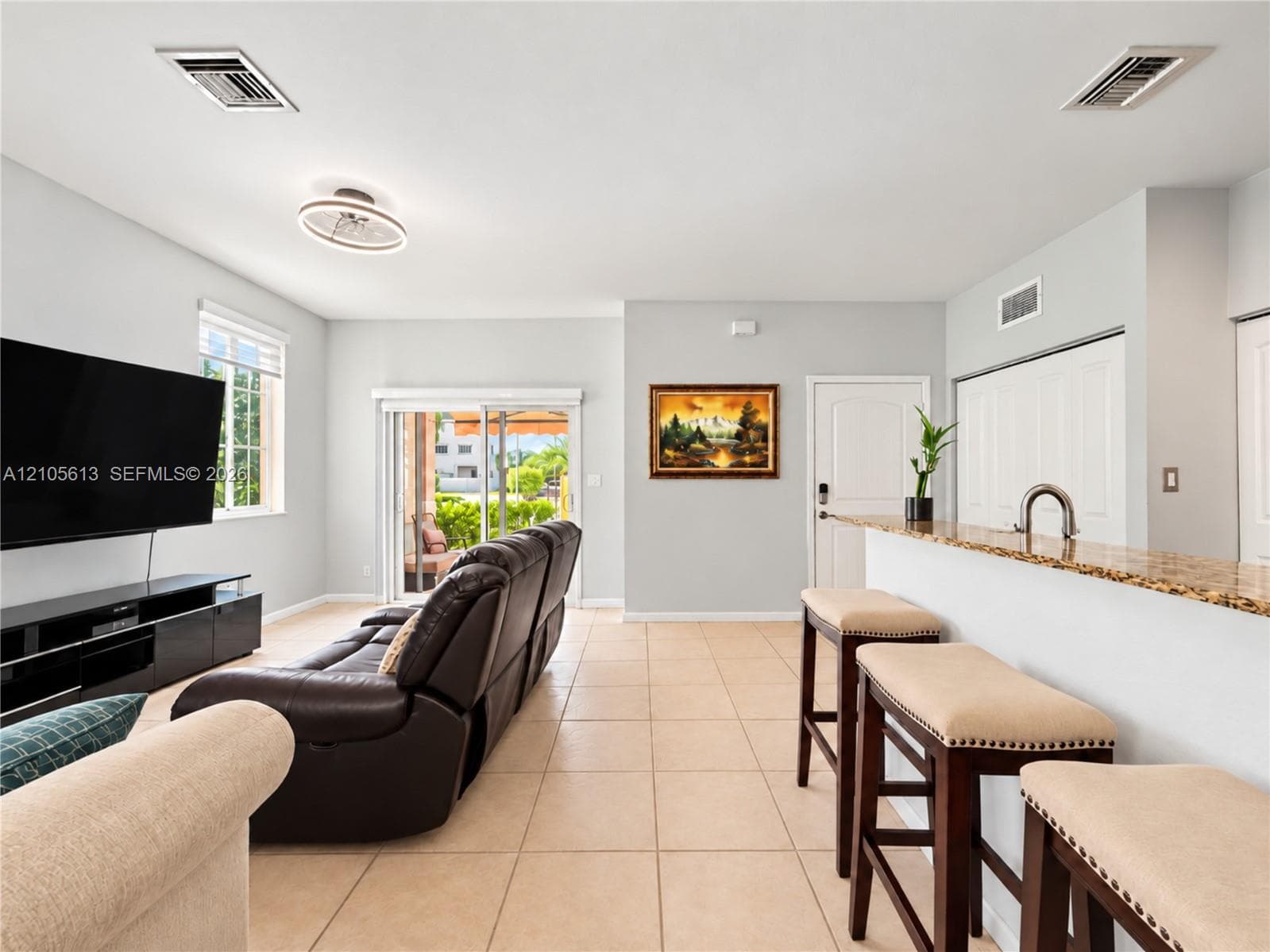 Open-concept interior featuring light-toned tile flooring, a stone-surfaced breakfast bar with a metallic faucet, sliding glass doors, and a modern circular flush-mount light fixture