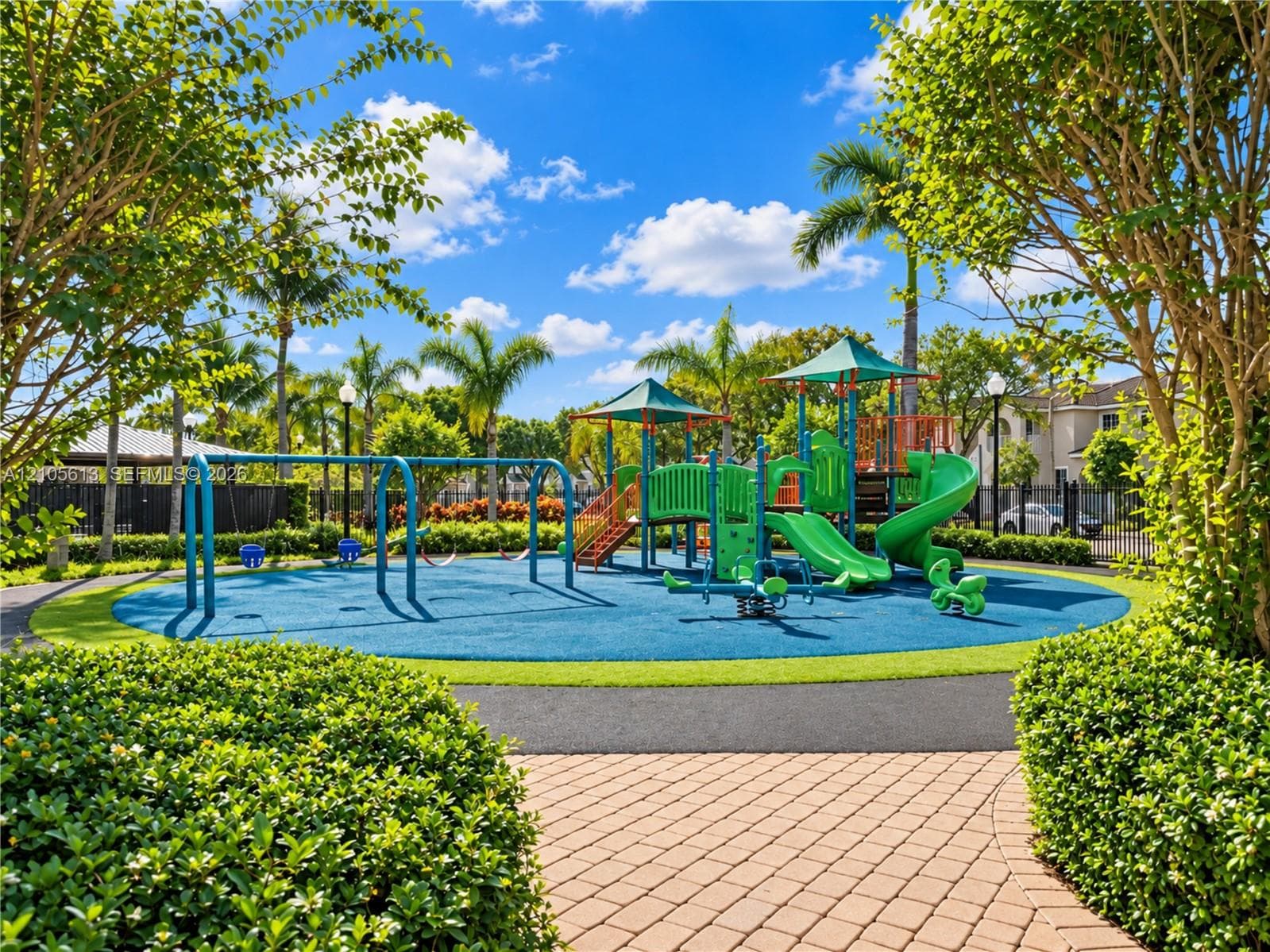 Outdoor recreation area featuring a multi-component play structure with slides, integrated swing set, rubberized play surface, and paved stone walkways surrounded by mature landscaping