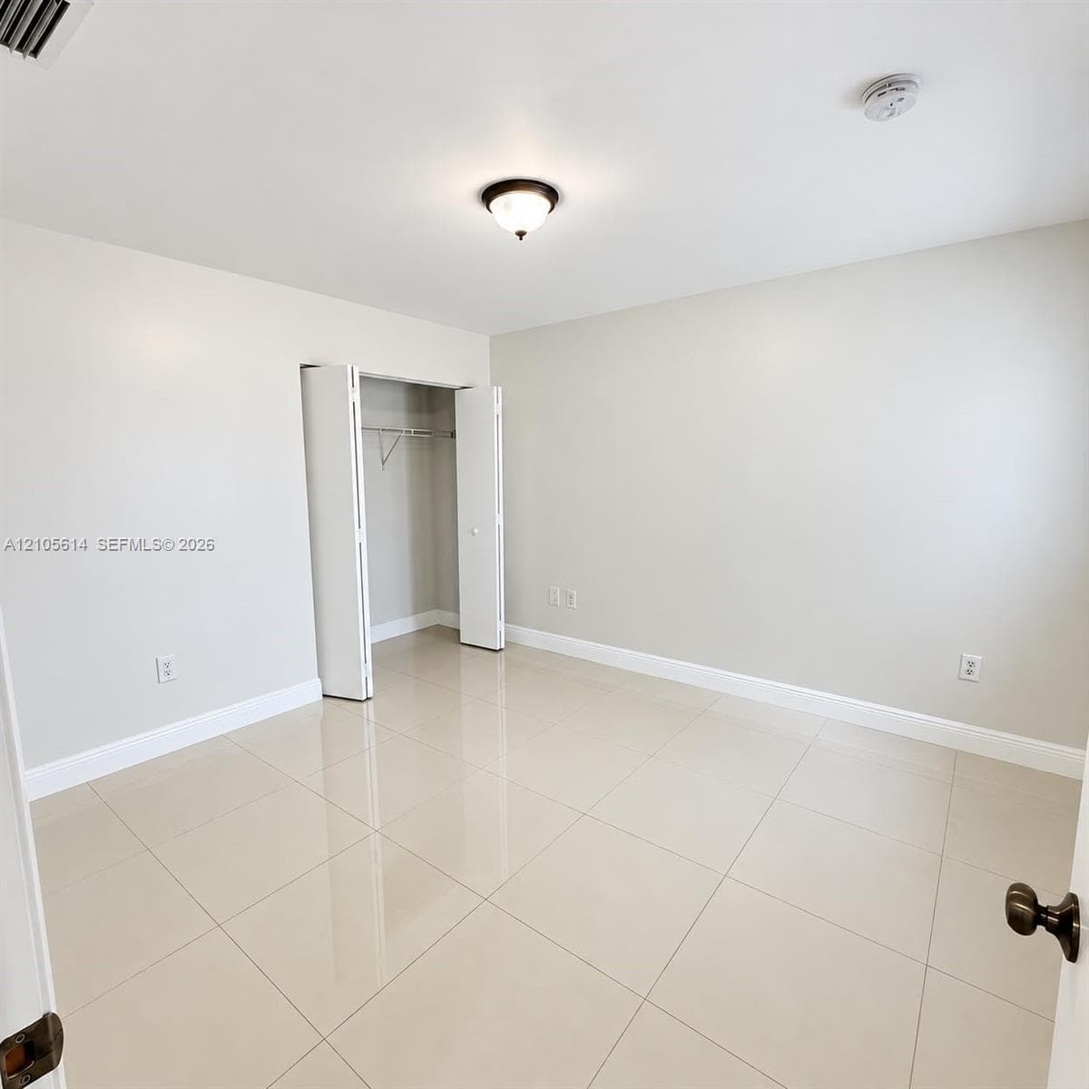 Neutral-toned room featuring large-format tile flooring, a walk-in closet with double-hinged doors, and a central ceiling-mounted light fixture