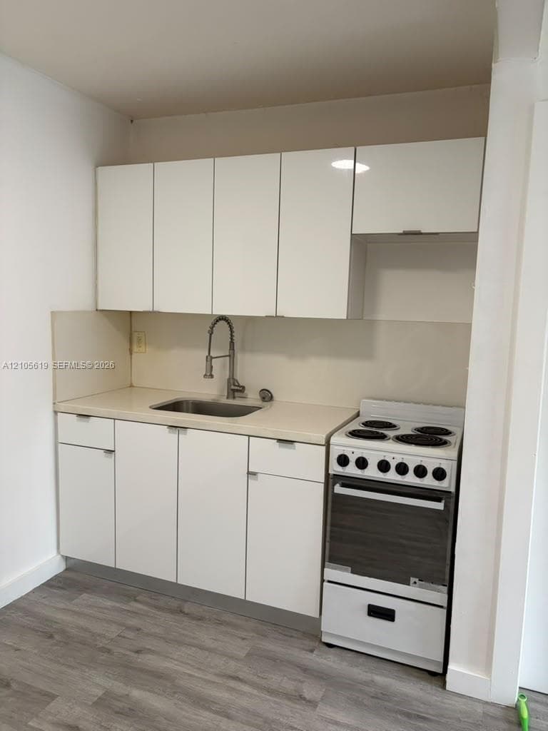 Kitchenette featuring high-gloss white flat-panel cabinetry, solid-surface countertop, stainless steel drop-in sink with a high-arch gooseneck faucet, four-burner electric range, and wood-finish flooring