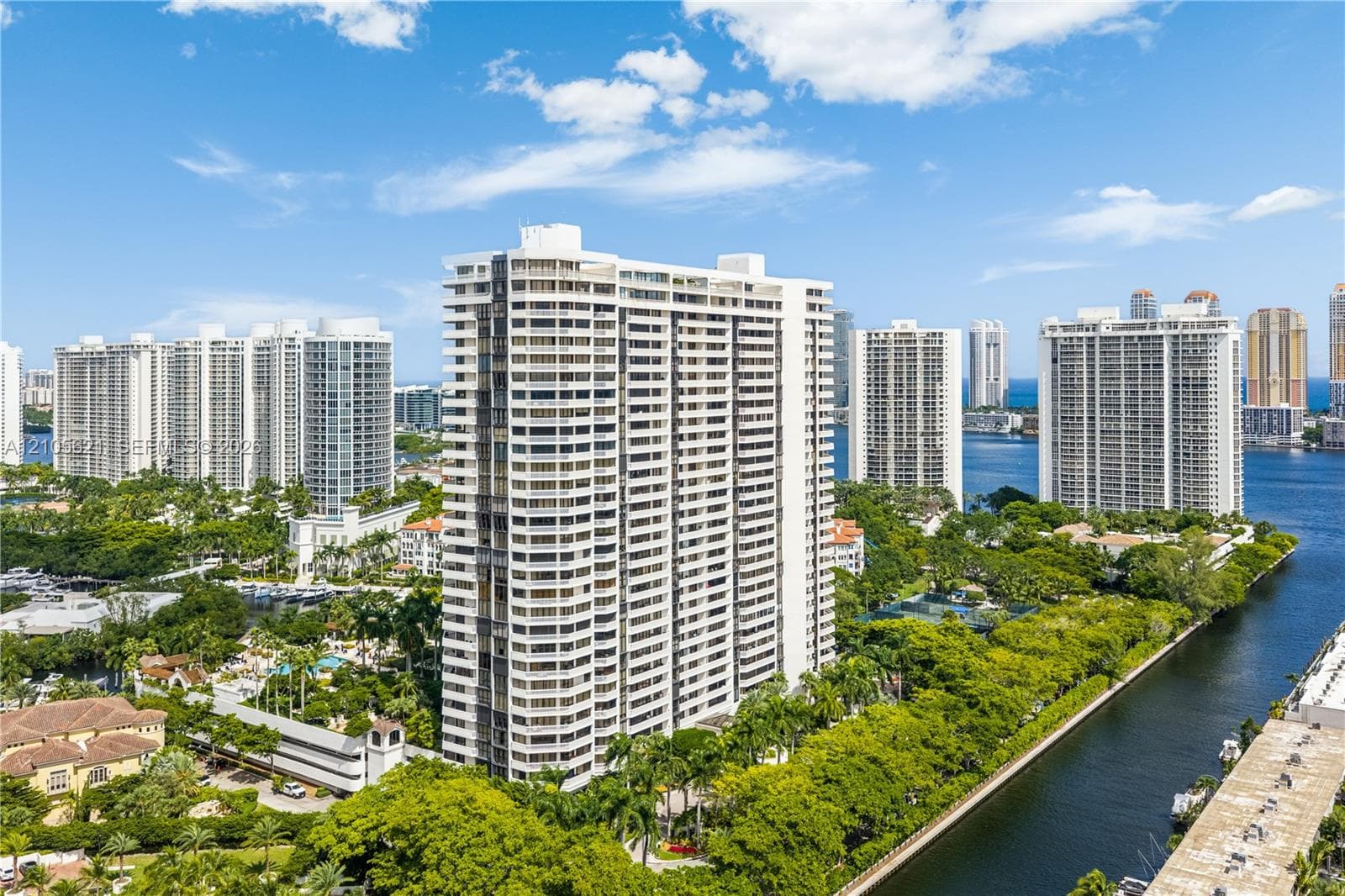 High-rise residential building featuring white concrete facade, recessed private balconies, expansive waterfront positioning, and surrounding tropical landscaping