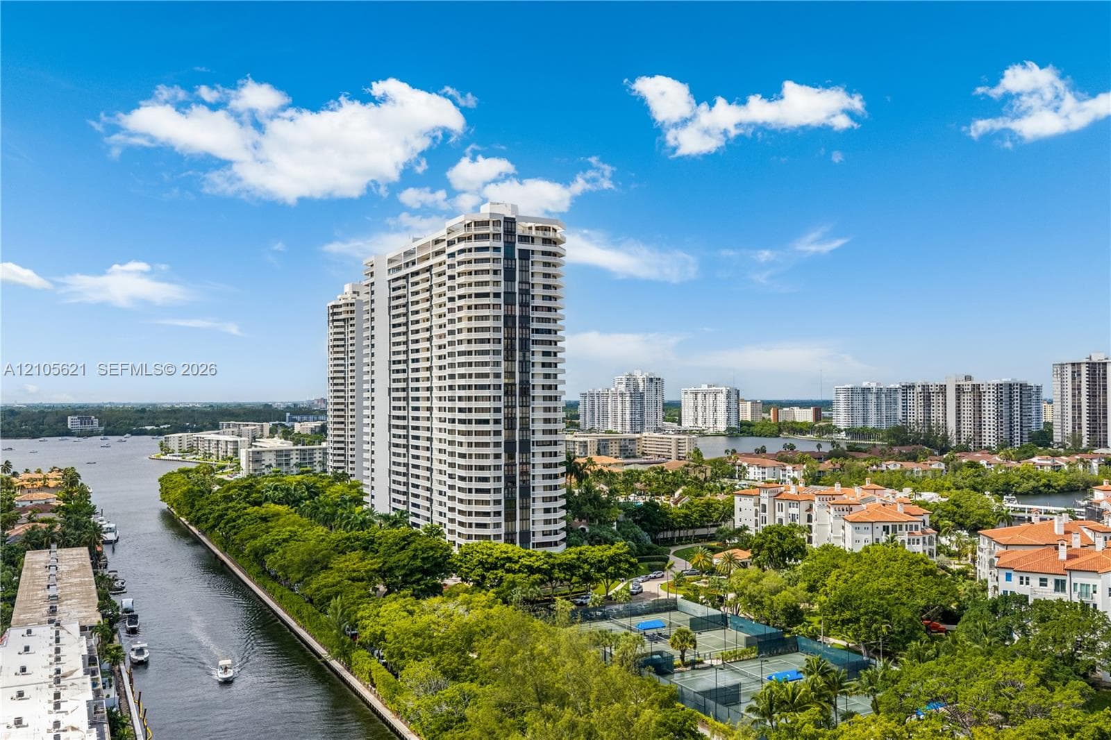 High-rise residential structure featuring tiered balconies, water frontage, tennis courts, and mature landscaping