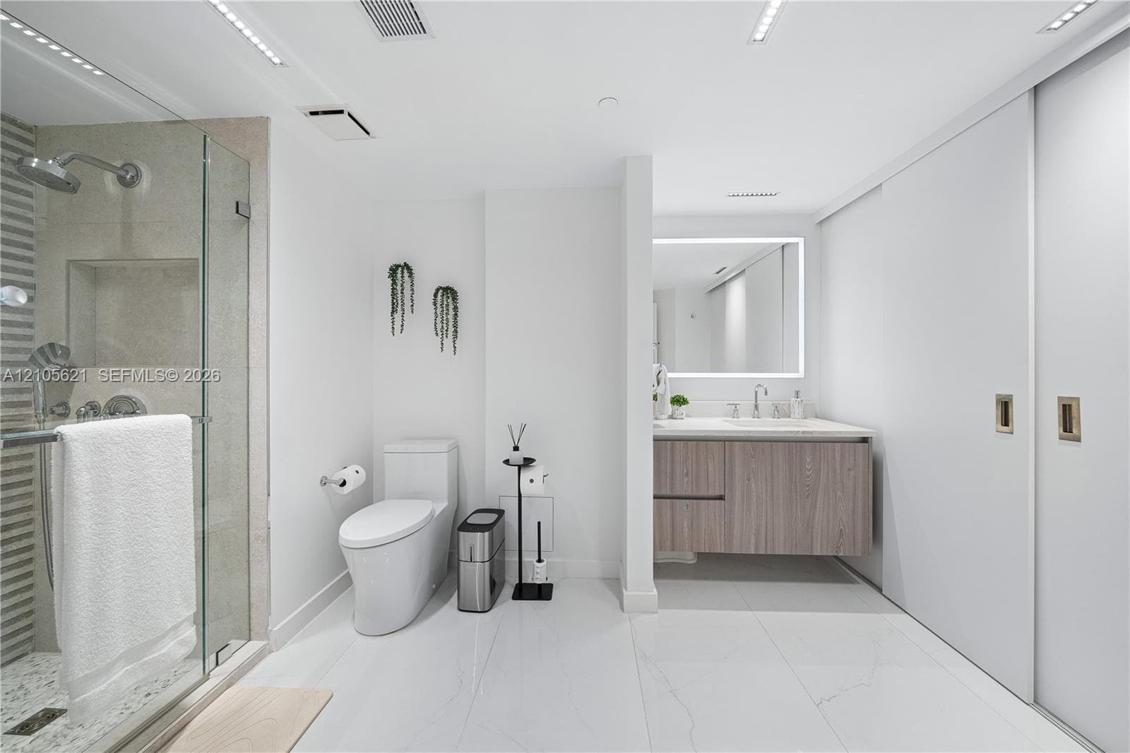 Modern bathroom featuring a glass-enclosed shower, wall-mounted wood-finish vanity with a light countertop, LED-backlit mirror, and white tile flooring