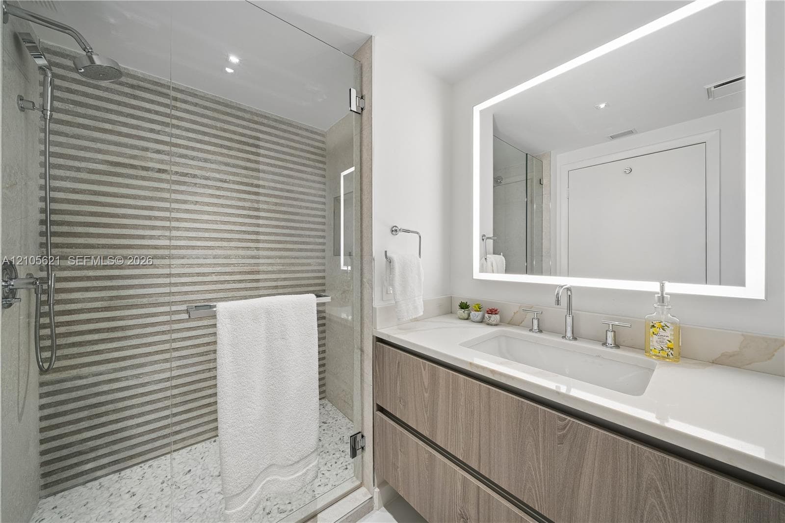 Floating vanity with wood-finish cabinetry, quartz countertop, integrated rectangular sink, wall-to-wall LED-backlit mirror, and walk-in shower with textured tile surround and pebble-style stone flooring