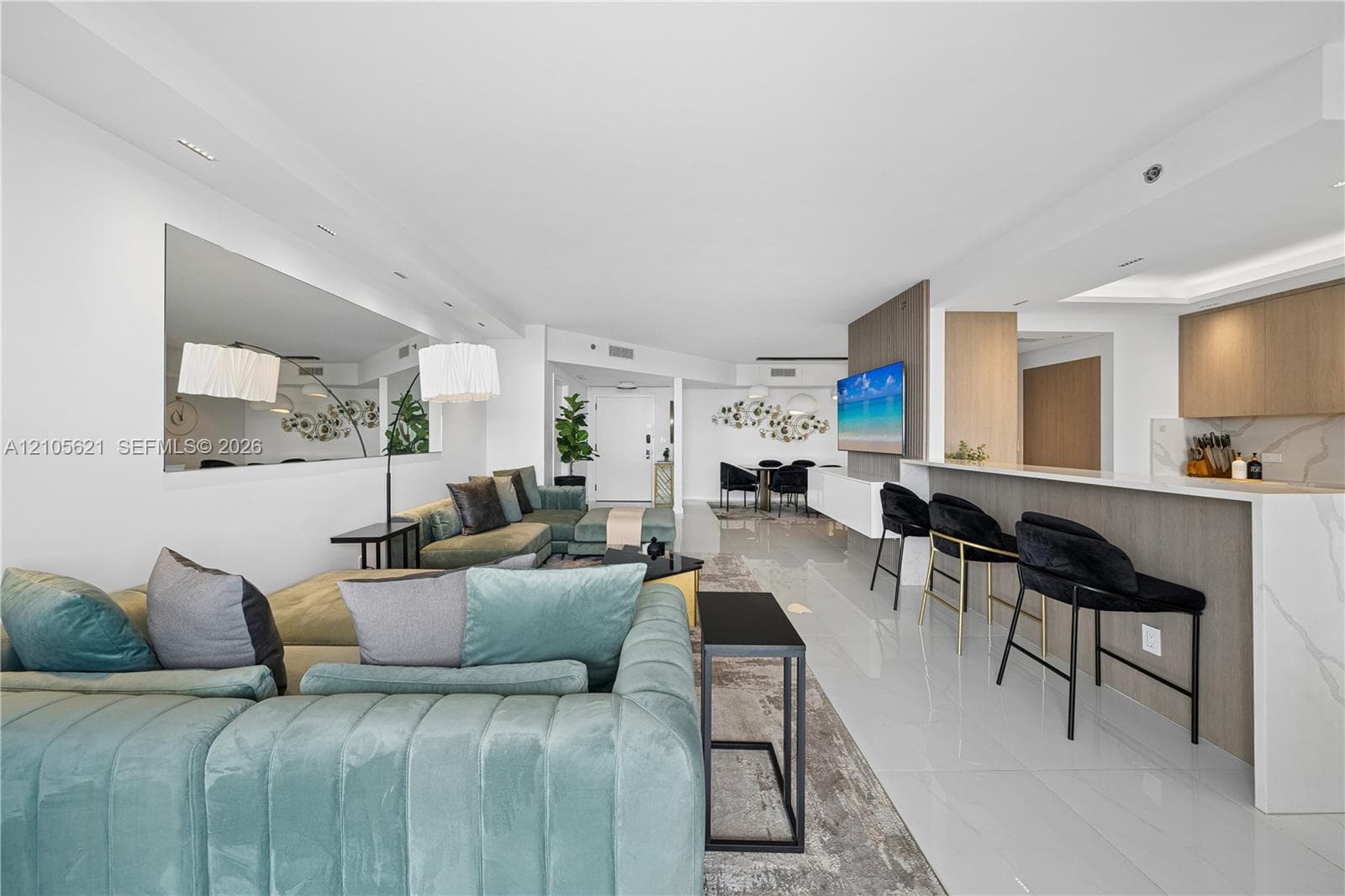 Open-concept living space featuring high-gloss tile flooring, recessed lighting, expansive wall-mounted mirror, wood-finish cabinetry, and kitchen peninsula with a stone-surfaced countertop