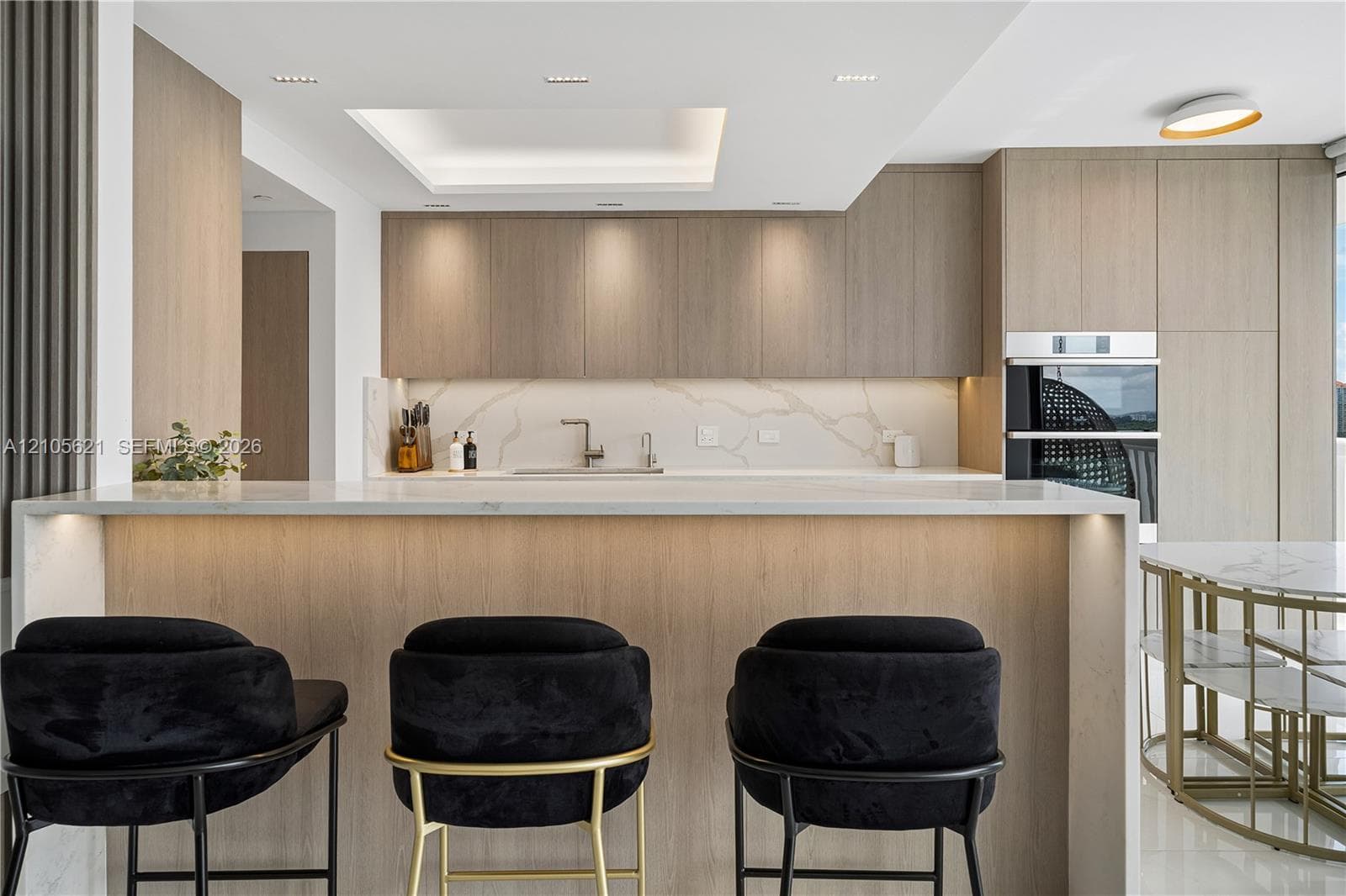 Modern kitchen featuring wood-finish cabinetry, waterfall-edge stone countertop, integrated double oven, and recessed tray ceiling lighting