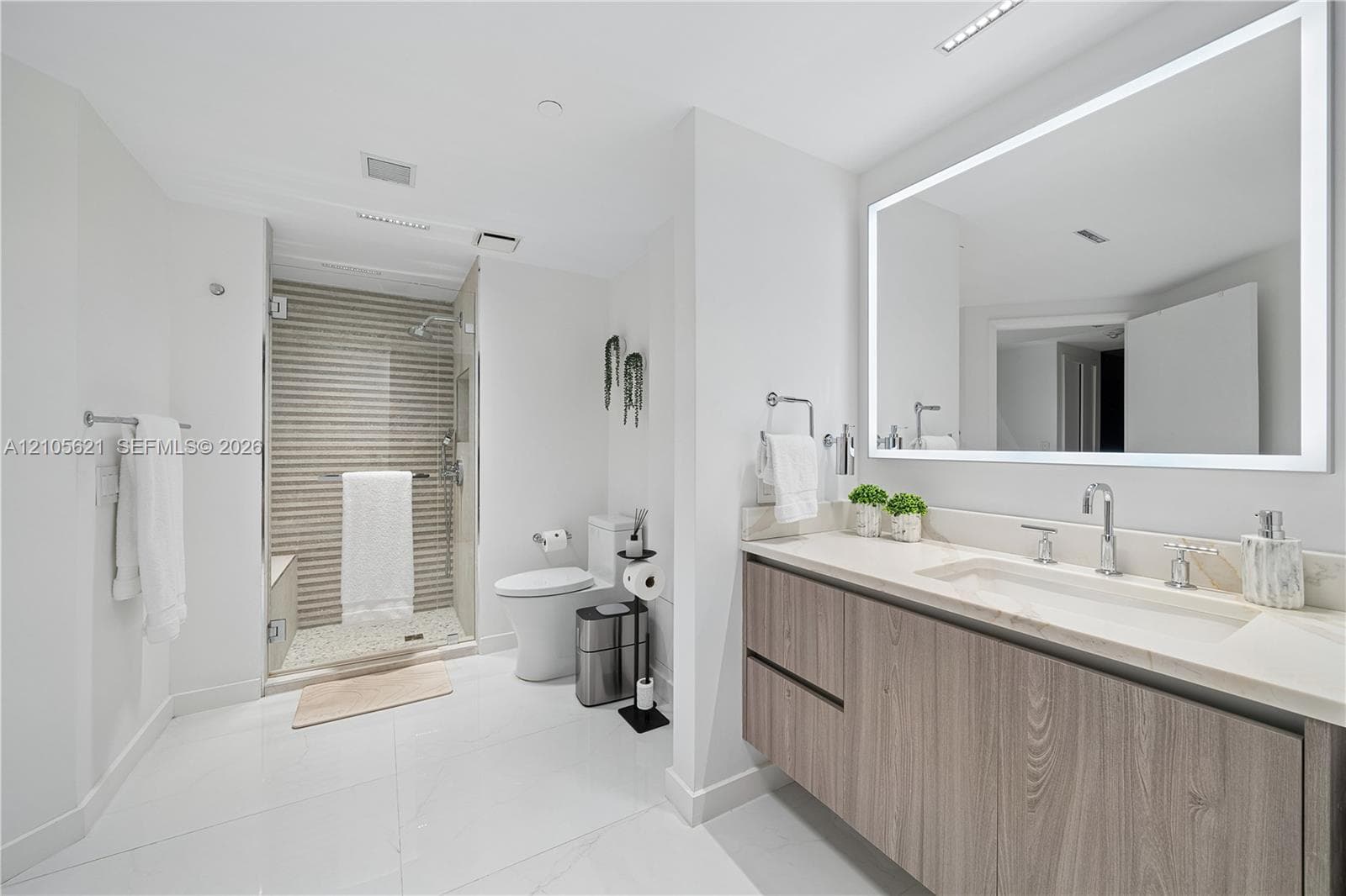 Floating wood-finish vanity with an integrated countertop, large rectangular mirror with perimeter integrated lighting, walk-in shower with stone tile accent wall, and polished tile flooring