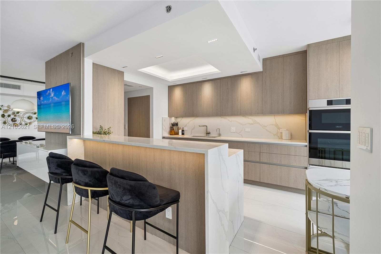 Kitchen featuring a waterfall-edge island with stone-finish countertops, flat-panel wood-finish cabinetry, recessed ceiling lighting, and built-in double wall ovens