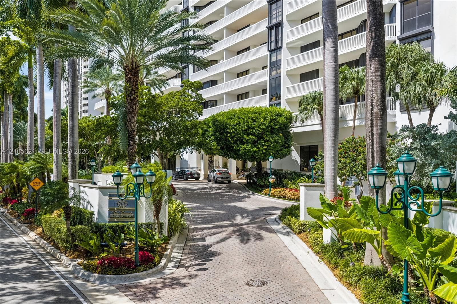 Multi-story residential structure featuring paved driveway, mature palm trees, tiered balconies with white railings, and manicured landscaping with integrated lantern-style light fixtures