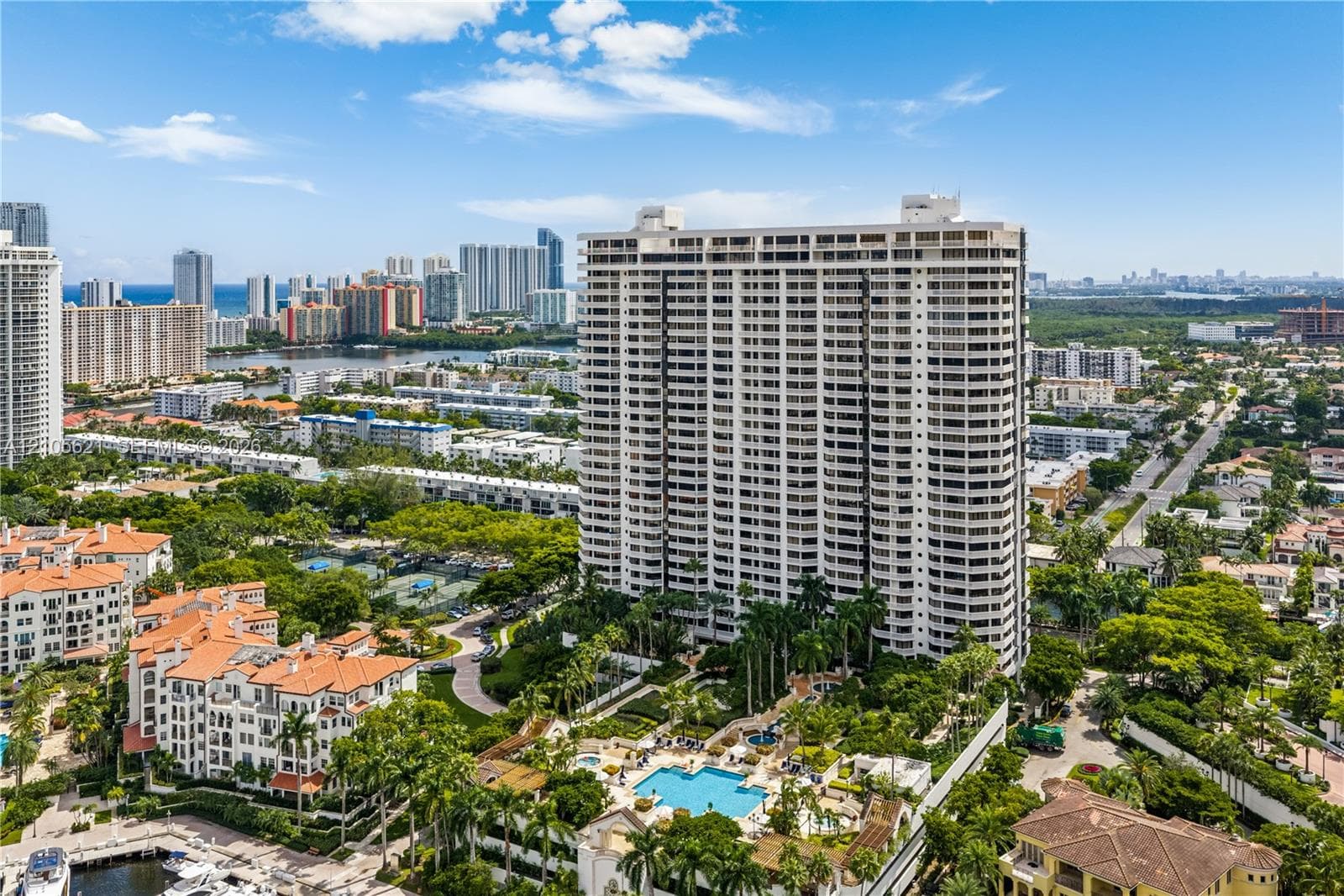 High-rise residential tower featuring wrap-around balconies, an outdoor swimming pool, mature palm trees, and landscaped grounds