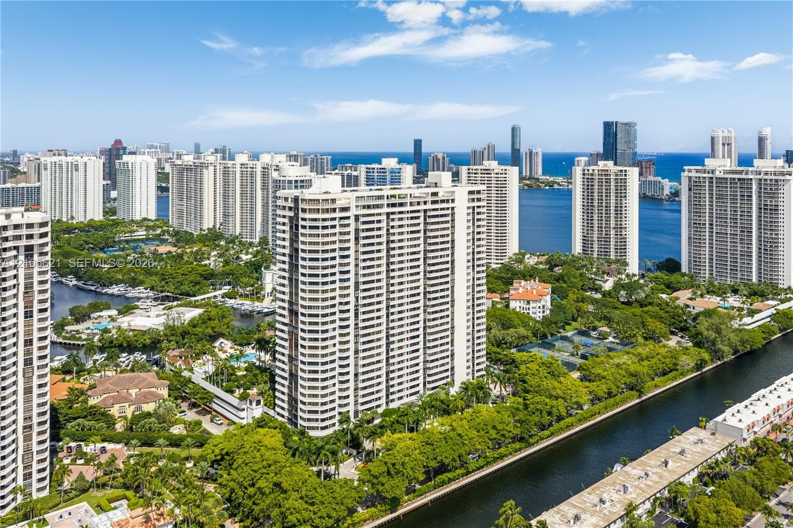 High-rise residential tower featuring balconies, expansive water frontage, surrounding mature landscaping, and proximity to the coastline