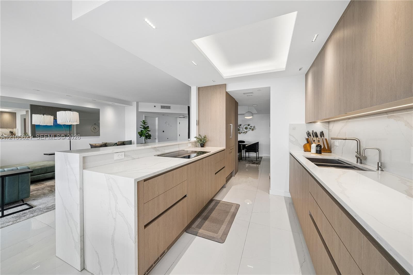 Contemporary kitchen featuring flat-panel wood-finish cabinetry, white stone countertops with waterfall edge, integrated cooktop, recessed tray ceiling lighting, and large-format tile flooring