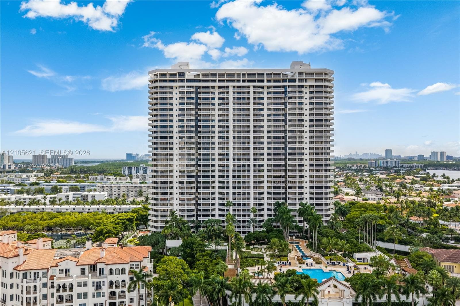 High-rise building exterior featuring continuous balconies, concrete structural detailing, ground-level swimming pool, extensive landscaping, and palm trees