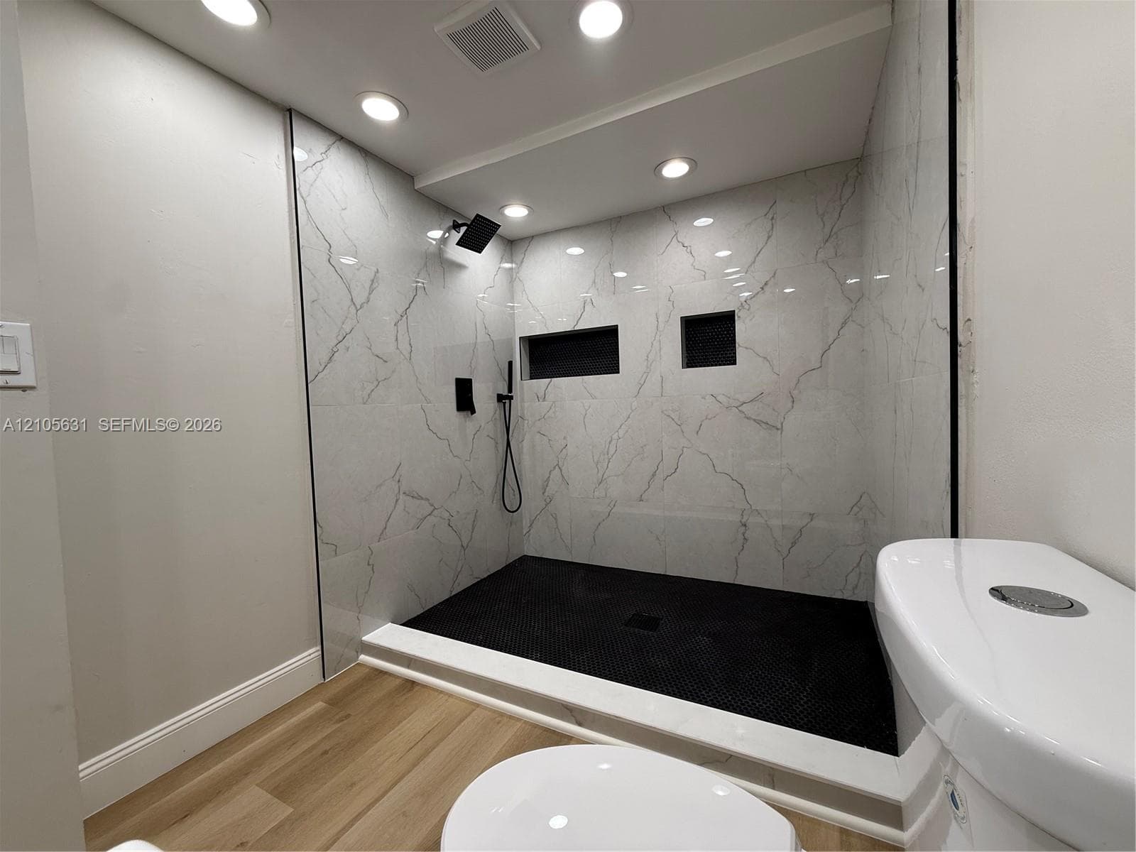 Walk-in shower featuring large-format veined tile surround, matte black rainfall showerhead, integrated wall niches, and black mosaic tile flooring, finished with wood-finish flooring