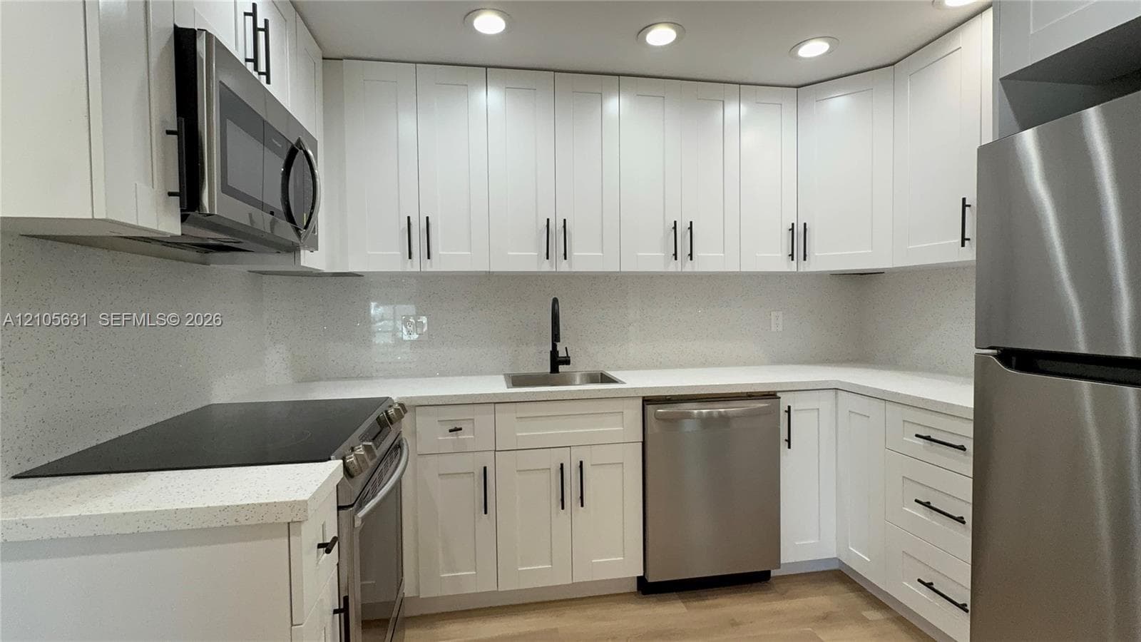 Kitchen featuring white Shaker-style cabinetry, light-toned composite countertops, a full-height solid surface backsplash, stainless steel appliances, and matte black hardware