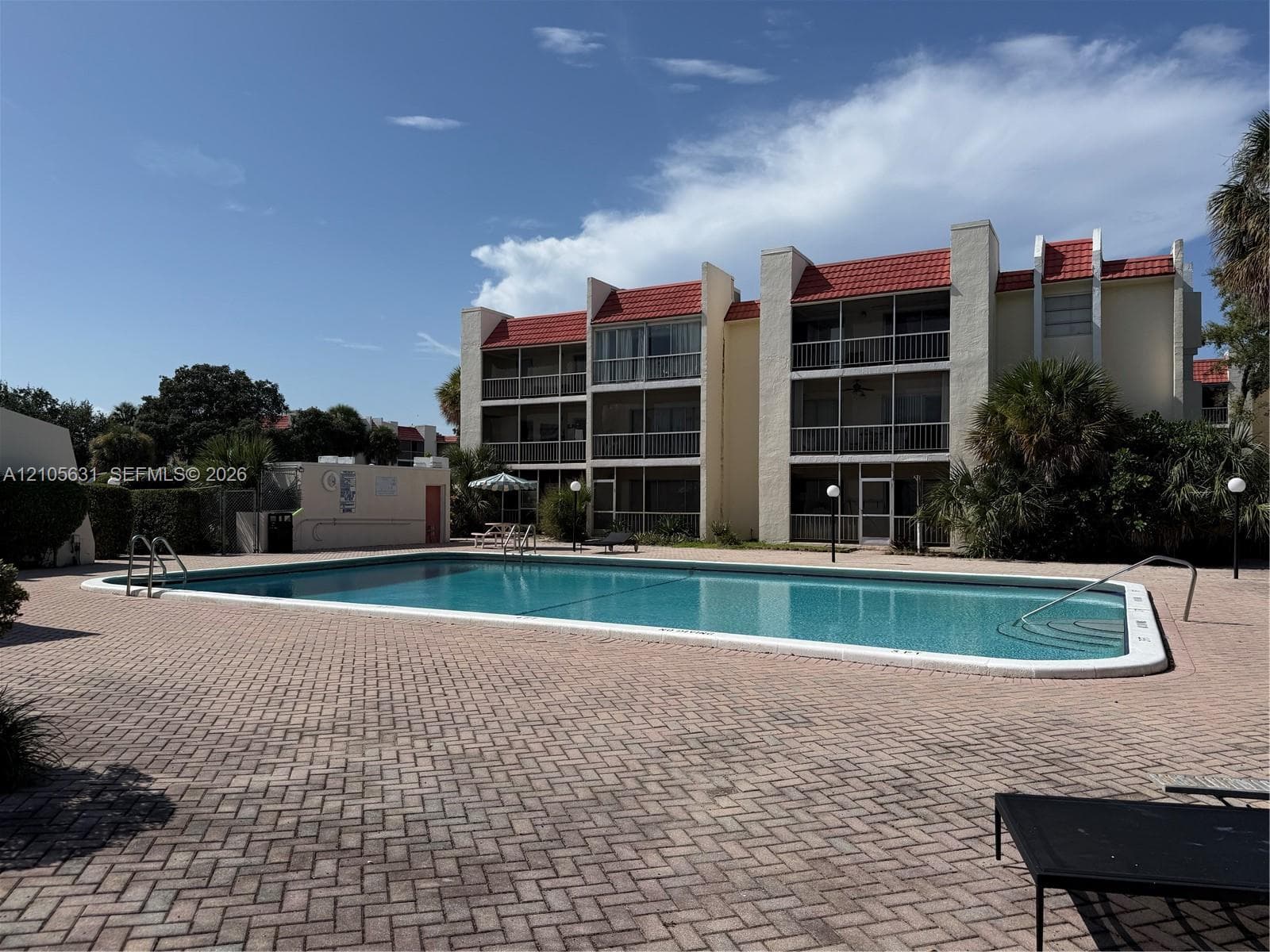 In-ground swimming pool with handrails, surrounded by a herringbone-patterned paver deck and multi-story building exterior featuring red tile roofing