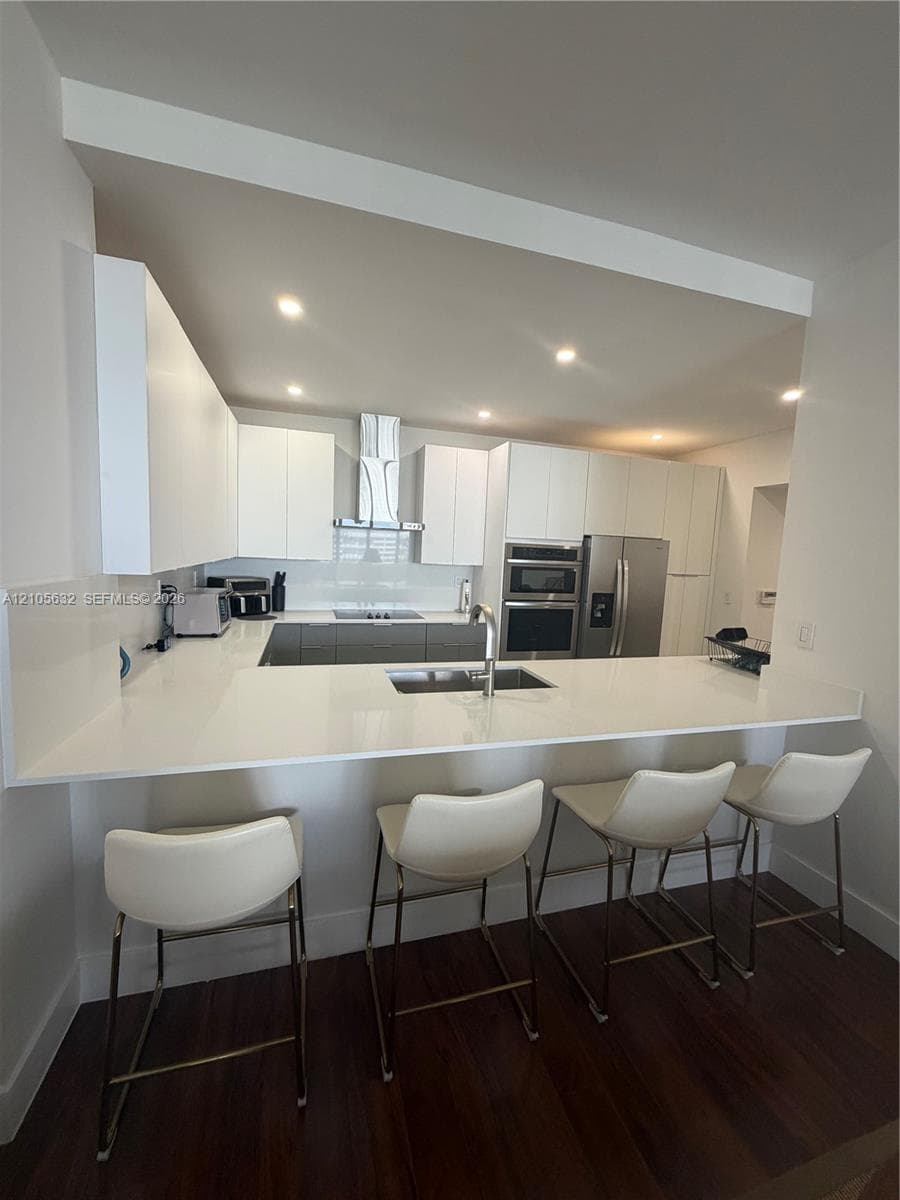 Modern kitchen featuring white flat-panel cabinetry, quartz countertops, stainless steel appliances, a stainless steel range hood, and wood-finish flooring