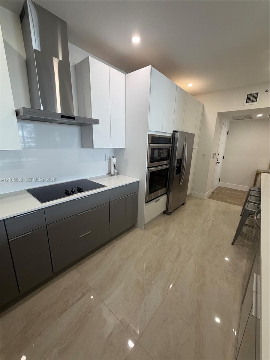Modern kitchen featuring two-tone cabinetry, stainless steel appliances, flat-panel wall ovens, stone-finish flooring, and a sleek stainless steel range hood
