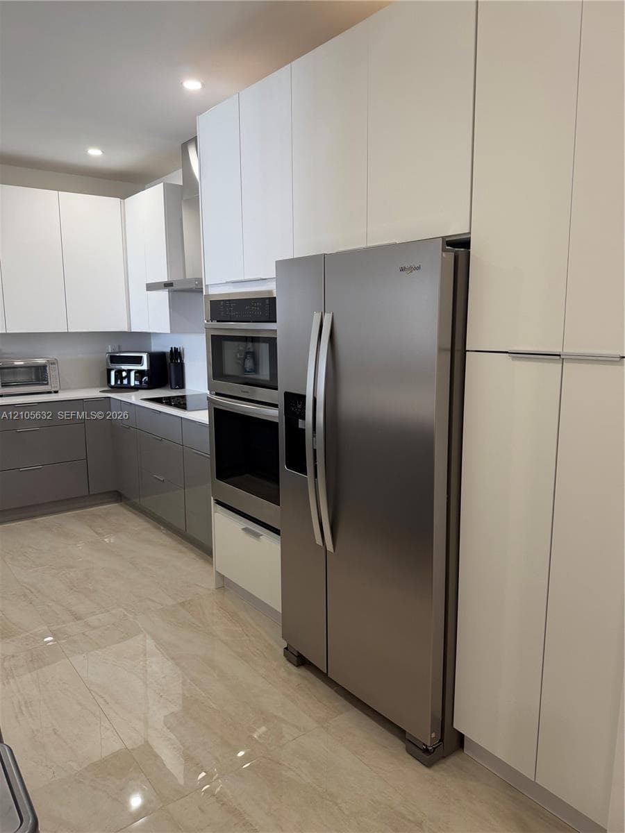 Modern kitchen featuring floor-to-ceiling white cabinetry, contrasting grey lower cabinetry, stone-finish tile flooring, and integrated stainless steel appliances including a built-in wall oven and microwave