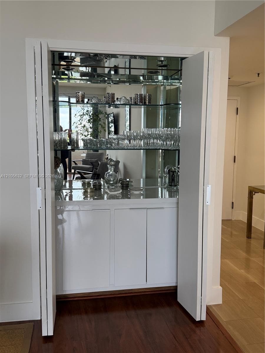 Built-in bar featuring white cabinetry, mirrored back paneling, and glass shelving enclosed by bifold doors