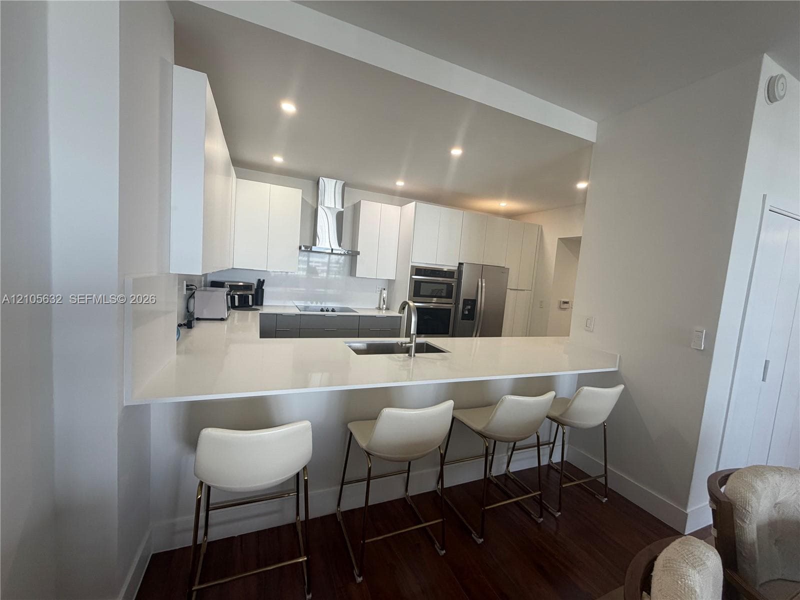 Kitchen featuring white flat-panel cabinetry, quartz countertops with a breakfast bar, recessed ceiling lighting, stainless steel built-in appliances, and wood-finish flooring